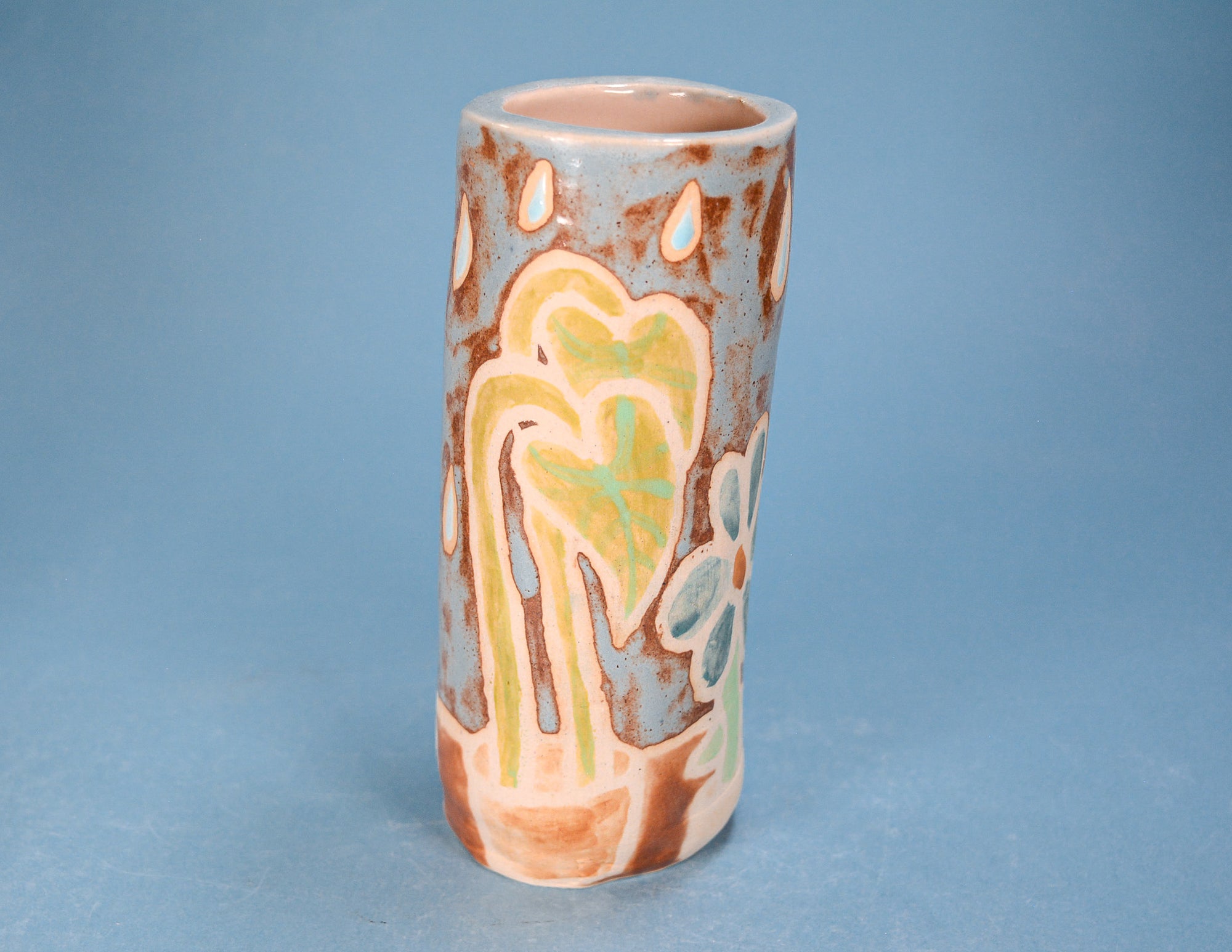 A handmade ceramic cylinder vase with an abstract, organic pattern in earthy brown, light blue, pale yellow, and mint green on a speckled off-white base, set against a solid blue background.