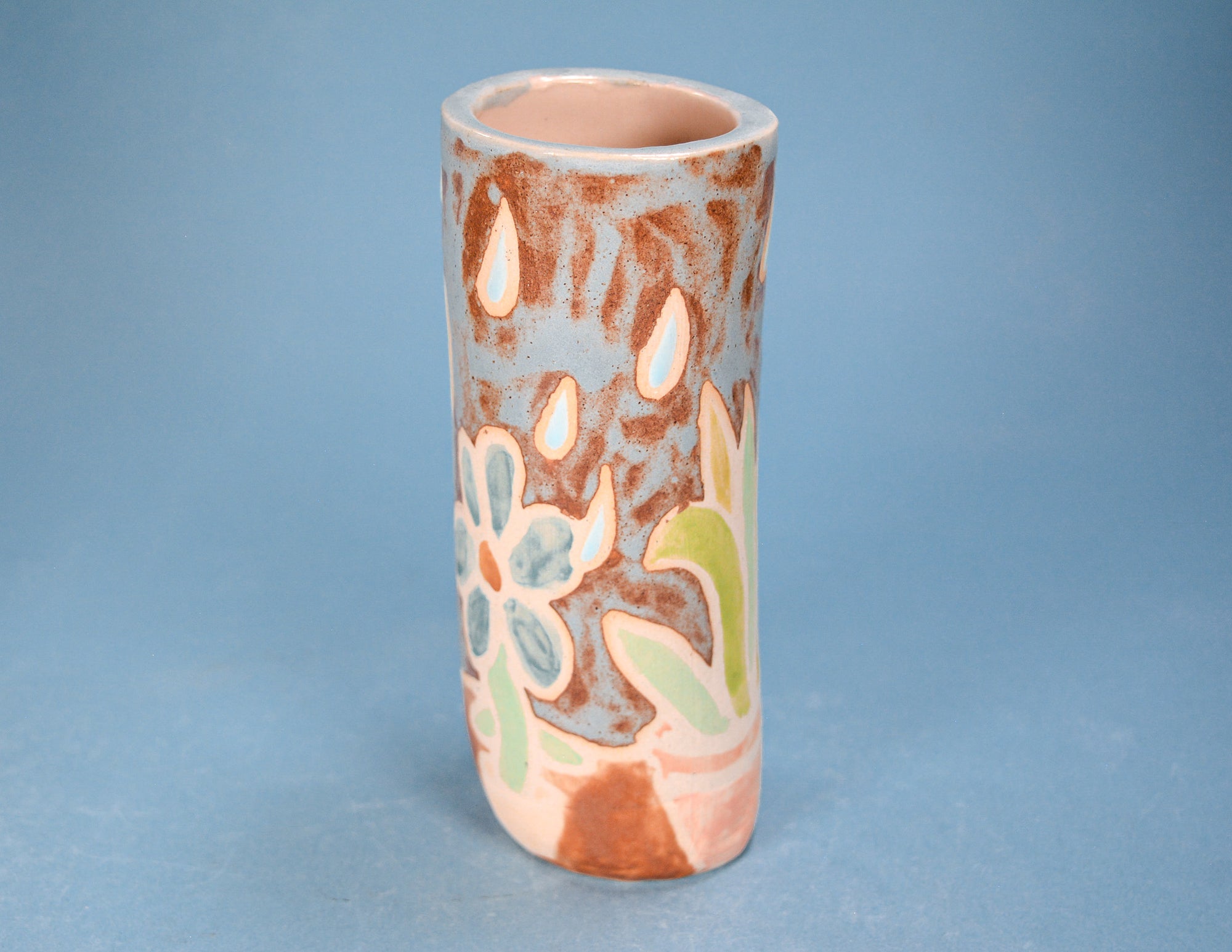 A small cylindrical ceramic vase with a flared rim, featuring a hand-painted floral and abstract pattern in terracotta brown, pale pink, light blue, and light green on a light blue-gray background, set against a solid blue backdrop.