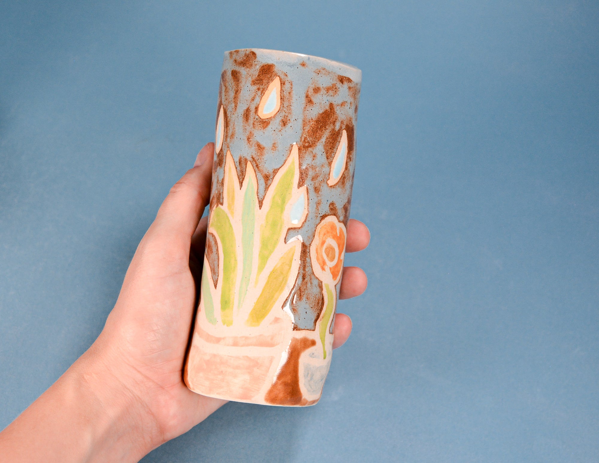 A hand holding a handmade ceramic cylinder vase with a blue-gray base, brown speckled glaze, and painted botanical motifs in green, peach, and cream, set against a solid blue background.
