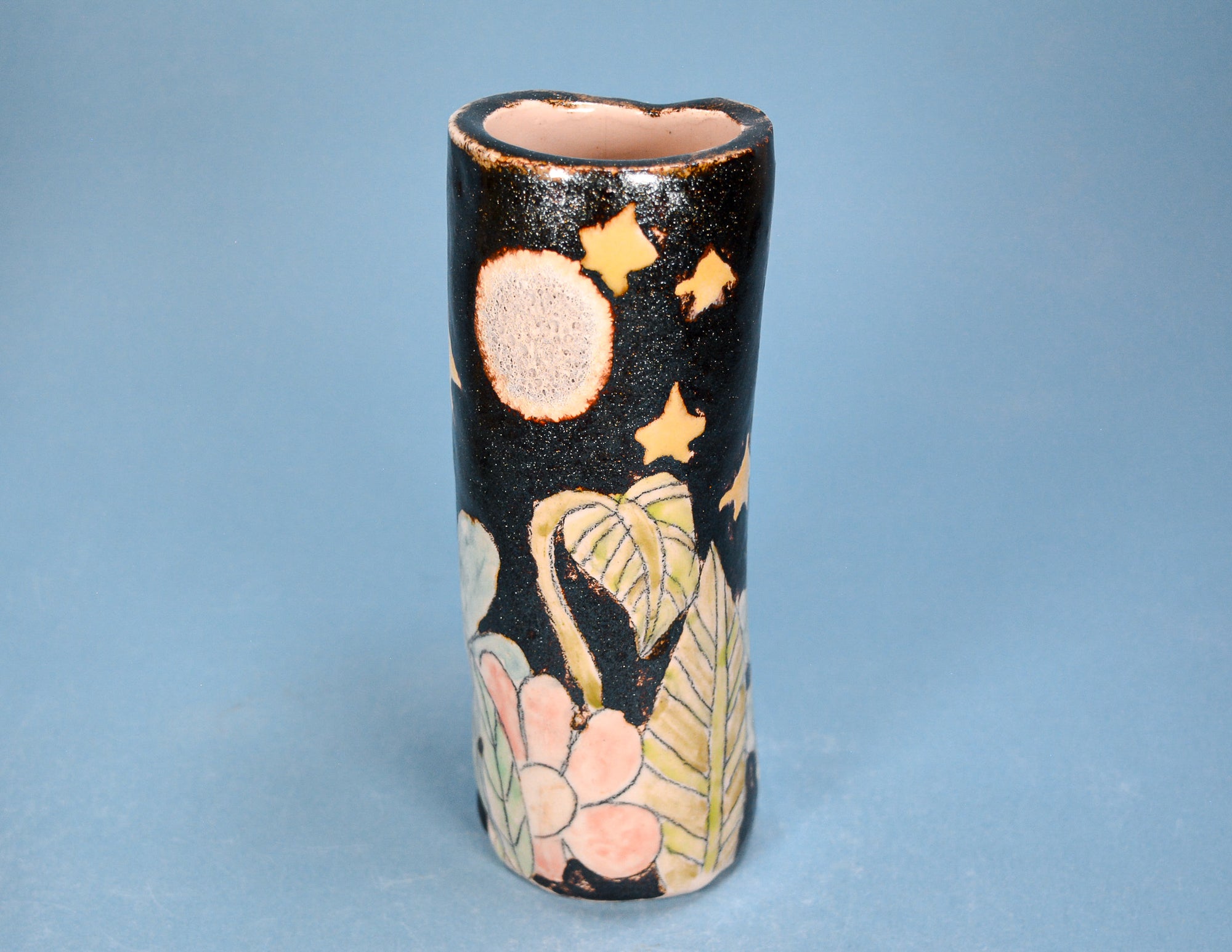 A small cylindrical ceramic vase with a black speckled glaze, decorated with gold stars and a pink and yellow floral pattern near the base, set against a solid blue background.