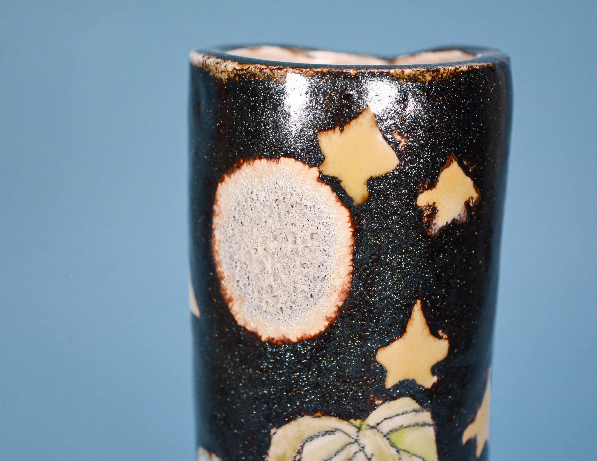 A close-up of a ceramic vase with a glossy black glaze decorated with cream-colored stars and a large textured cream moon, set against a solid blue background.