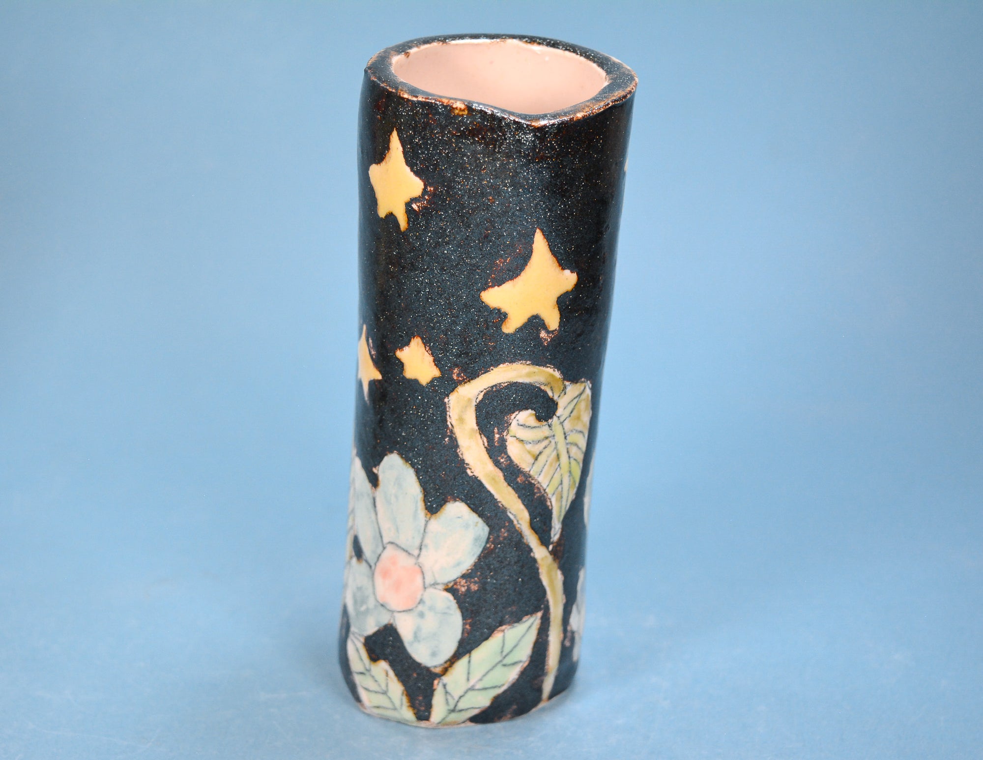 A small cylindrical ceramic vase with a black speckled glaze decorated with gold stars, a white flower with a pink center, and green leaves, set against a solid blue background.