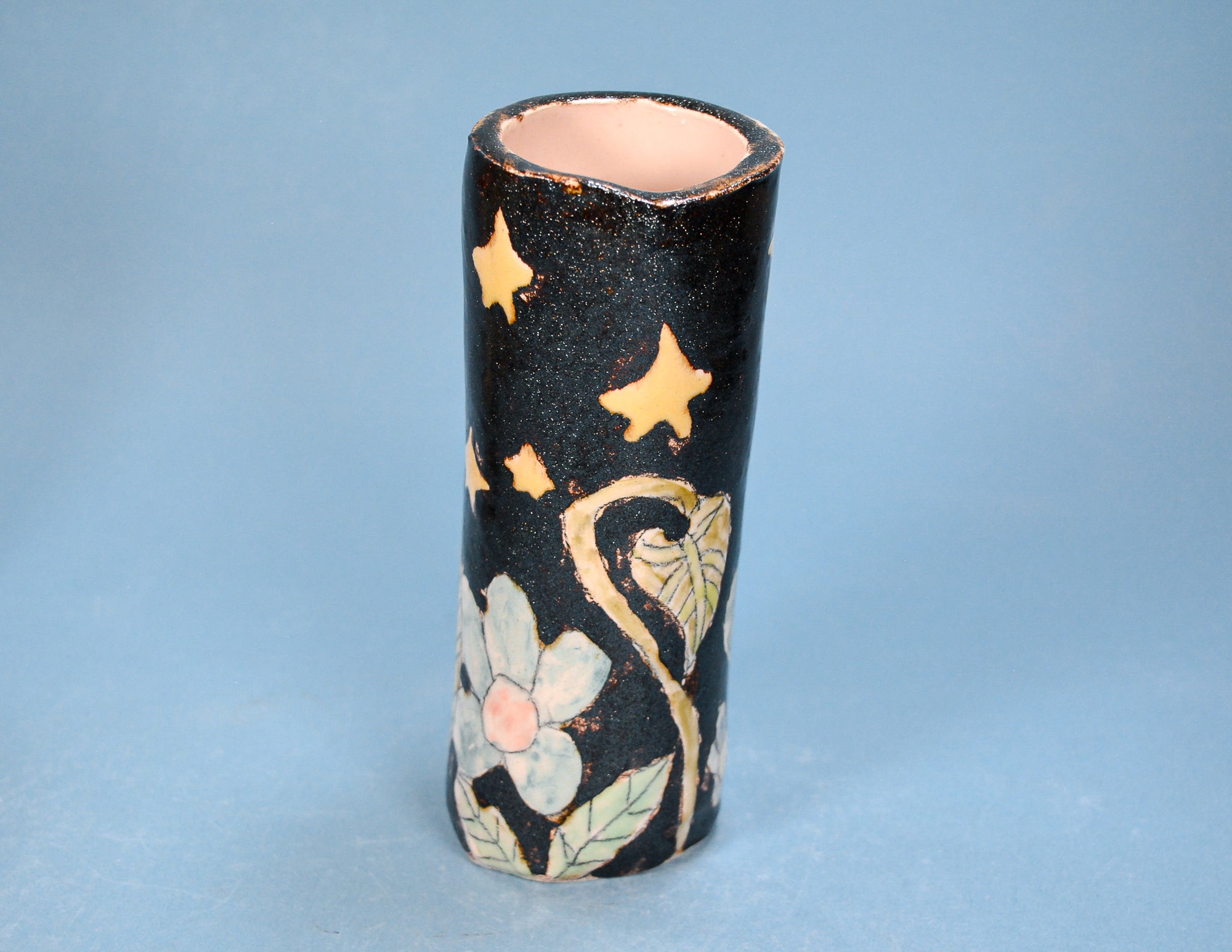 A small cylindrical ceramic vase with a glossy black glaze, decorated with gold stars, a gold crescent moon, and a white flower with a pink center near the base, set against a solid blue background.