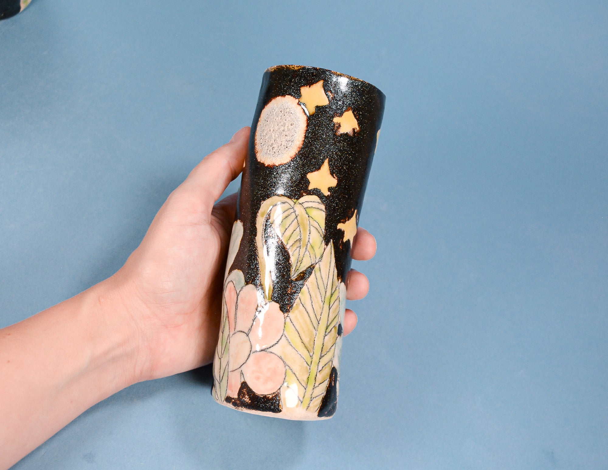 A hand holding a cylindrical ceramic vase decorated with a black sky motif featuring gold stars and a pale pink moon, alongside botanical patterns in pink and green on a light blue background.