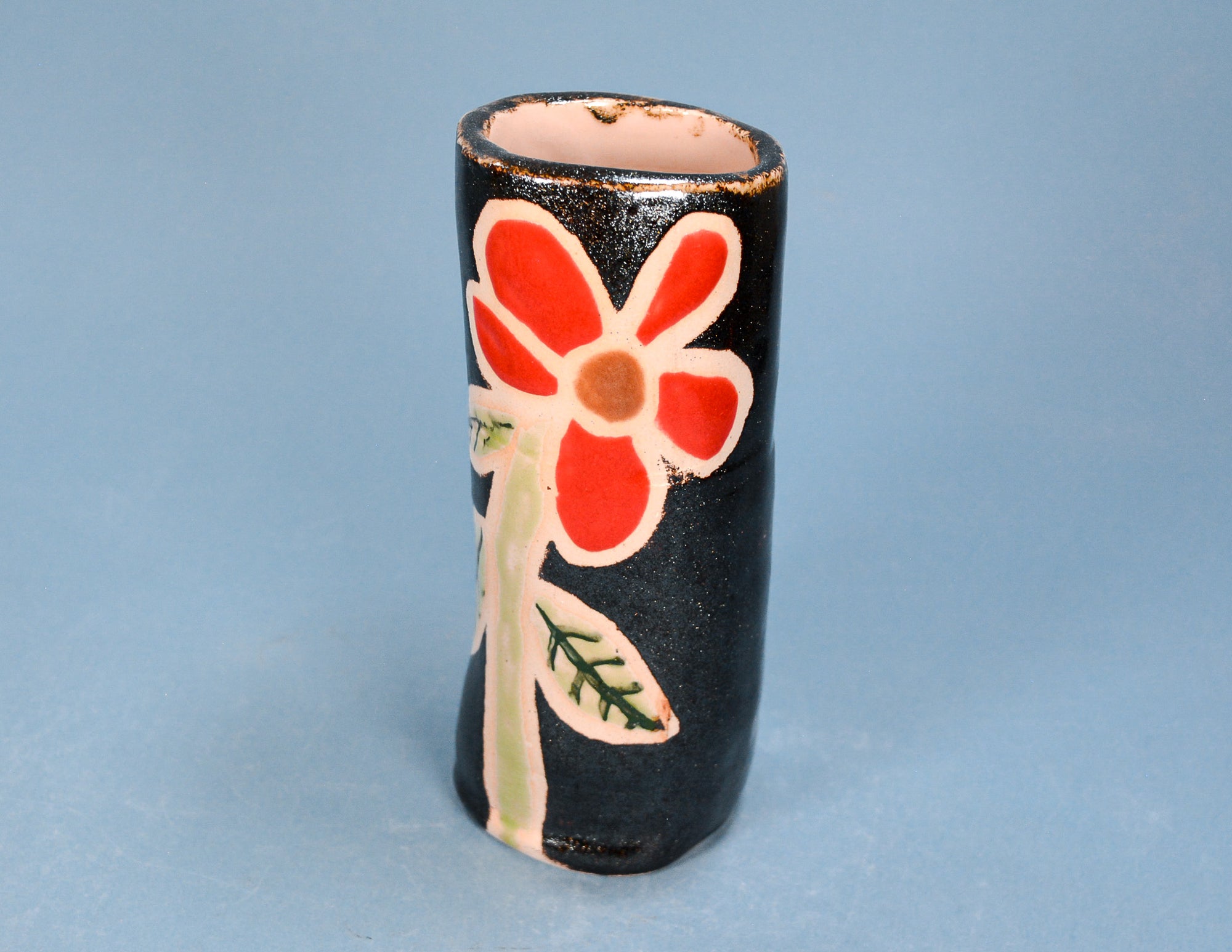 A small handmade ceramic vase with a glossy black body, featuring a hand-painted red flower with cream-colored petals and stem, and green leaves, set against a solid light blue background.