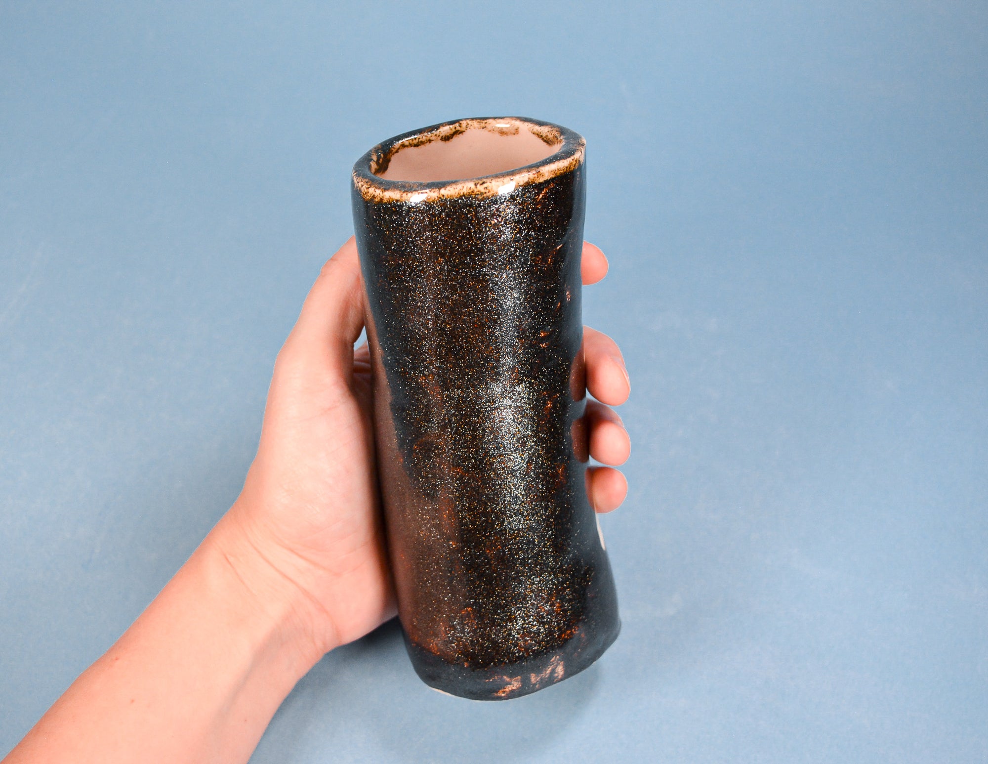 A hand holding a small, dark brown ceramic vase with a textured, metallic-looking glaze against a solid blue-gray background.