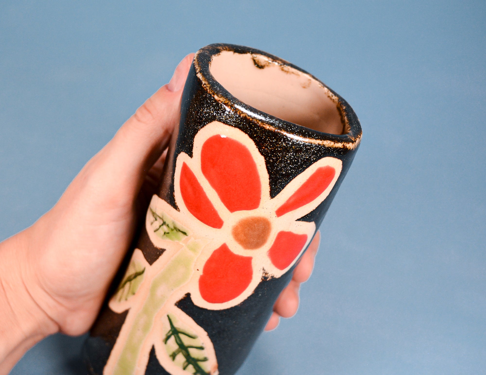 A hand holding a handmade ceramic cup or small vase with a black speckled glaze, decorated with a red flower and green leaves on a blue background.