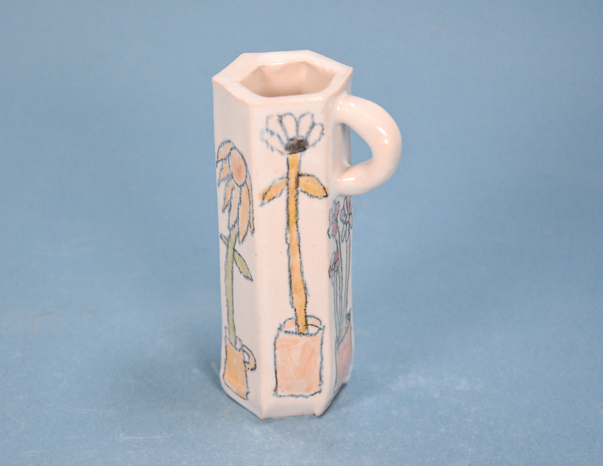 A small hexagonal ceramic vase with a handle, decorated with hand-painted dandelion illustrations in soft pastel colors, set against a solid light blue background.