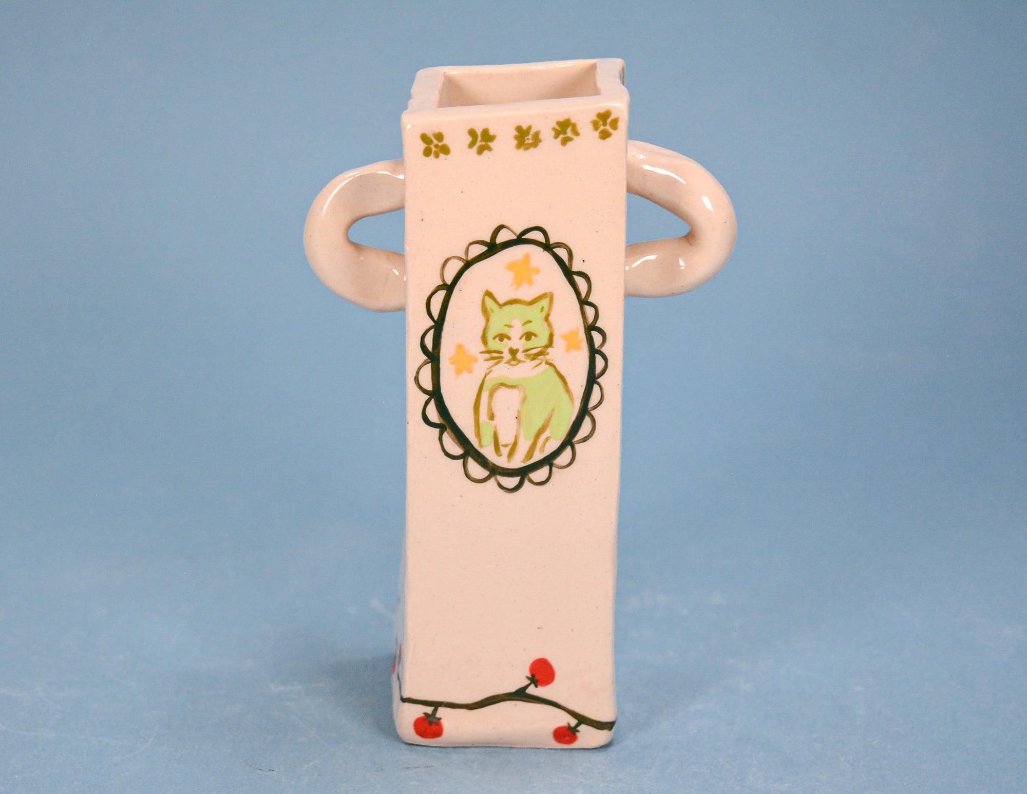 A pale pink ceramic vase with two loop handles, featuring a hand-painted green cat in an oval frame on the front, decorated with small yellow flowers near the top and red berries on the bottom, set against a solid blue background.