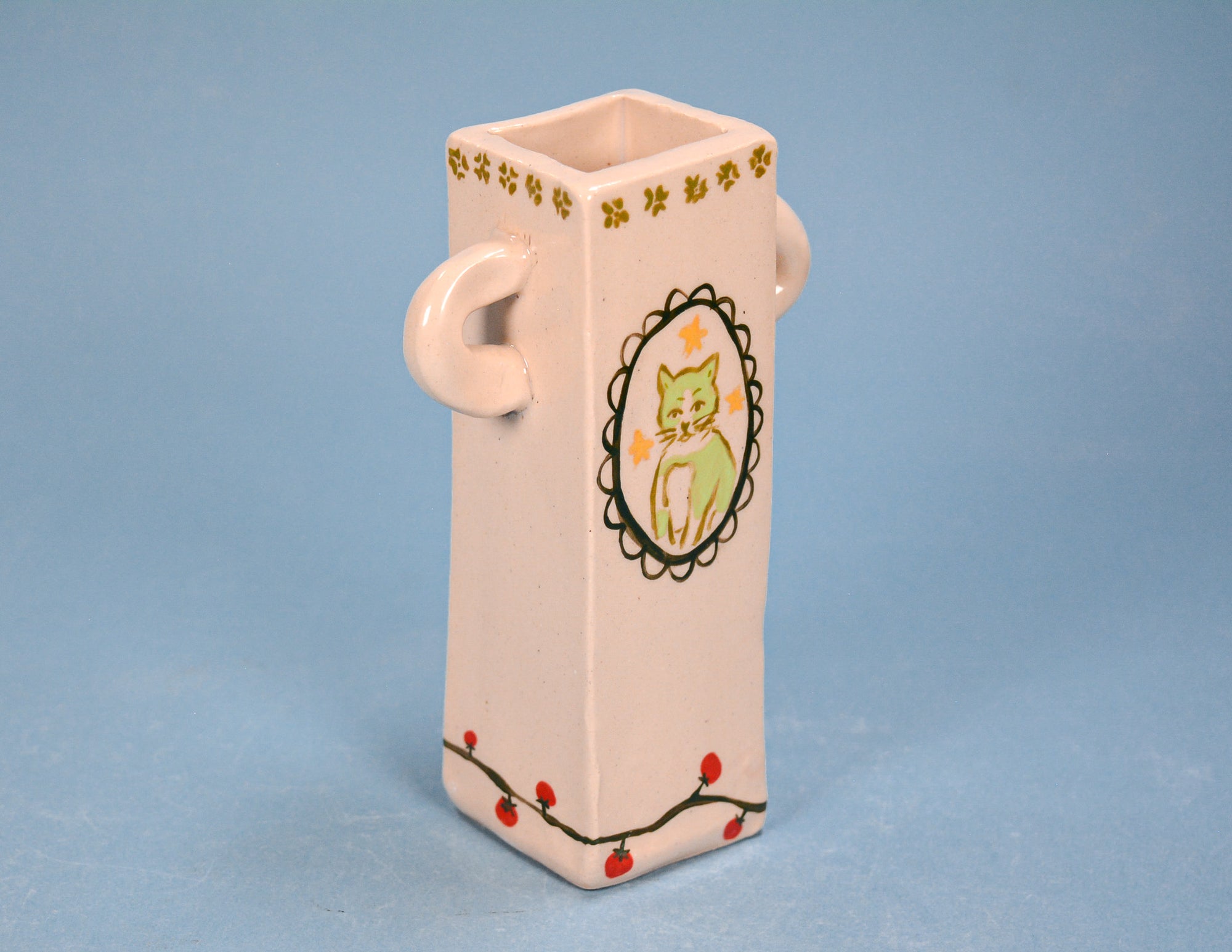 A small rectangular ceramic vase with side handles, decorated with a green cat illustration and floral details, set against a solid blue background.