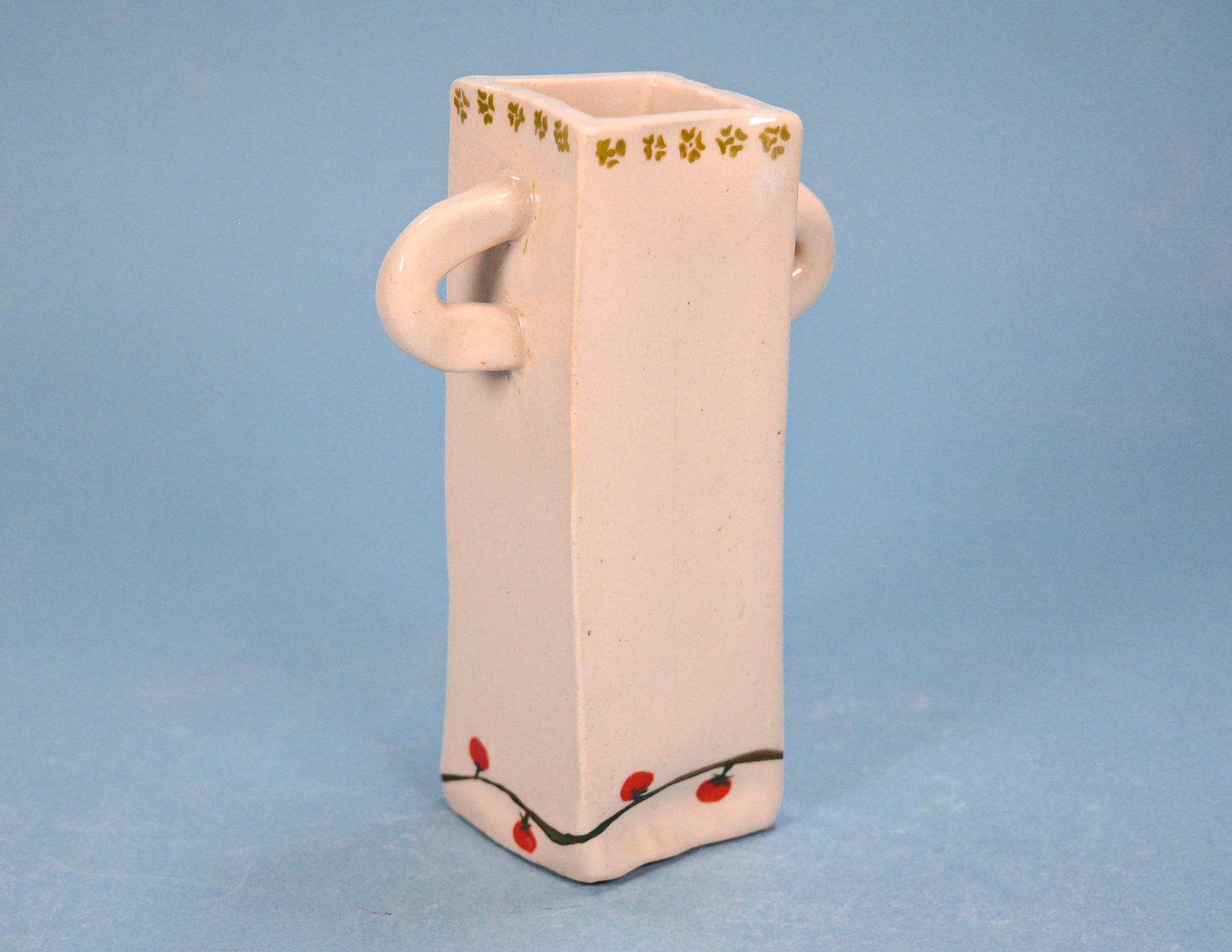 A light pink ceramic rectangular vase with two loop handles, decorated with a gold floral border on the rim and small red berries on a black vine near the base, set against a solid blue background.