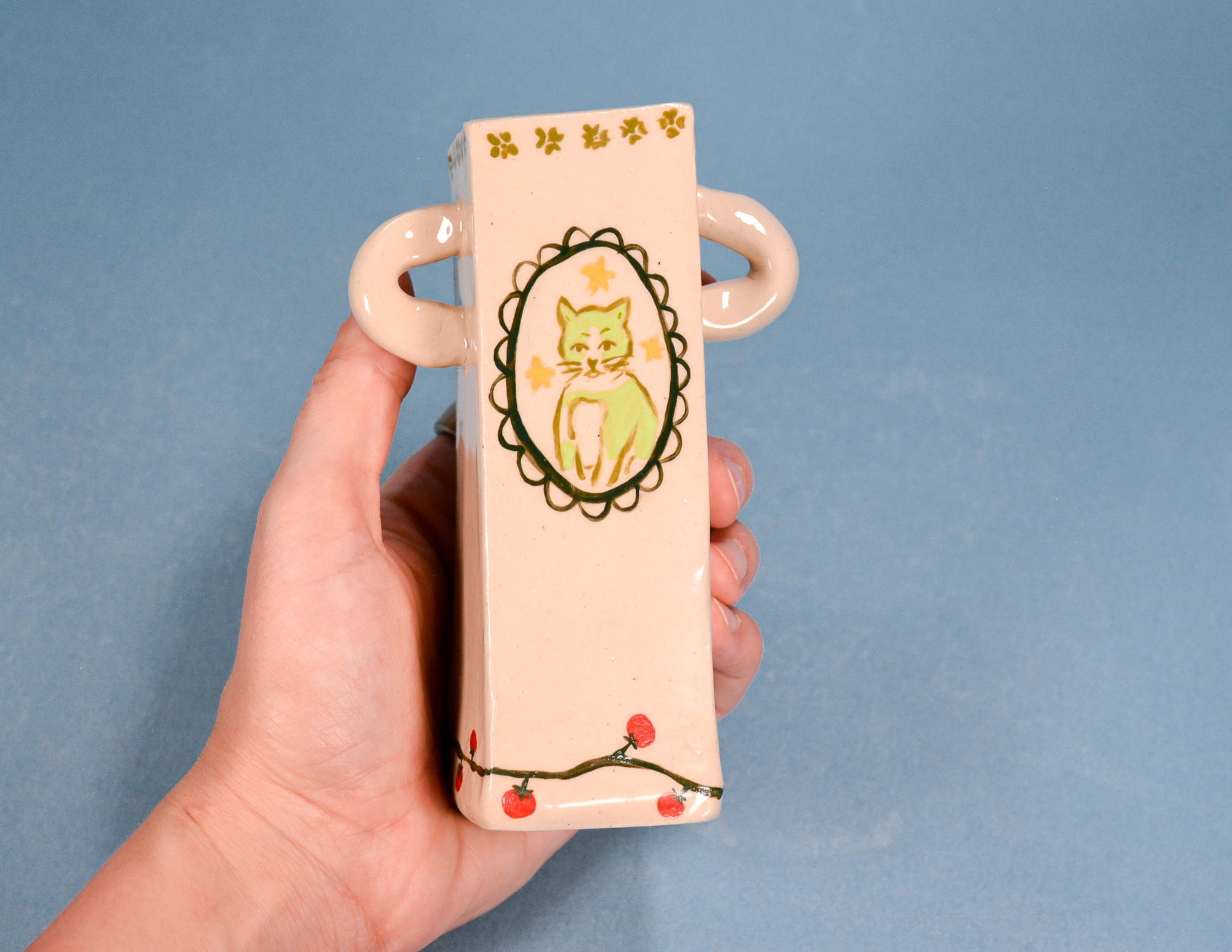 A hand holding a small rectangular ceramic vase with loop handles. The vase has a pale peach-pink glaze, decorated with a central illustration of a green cat inside a black scalloped oval frame, gold paw prints at the top, and a cherry branch with red berries at the bottom.