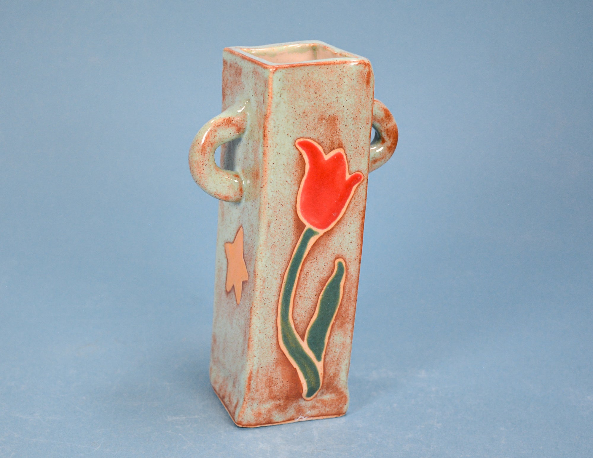 A tall rectangular ceramic vase with two loop handles on the sides, decorated with a red tulip and green stem motif on a speckled beige and orange-brown glaze, set against a solid blue-gray background.