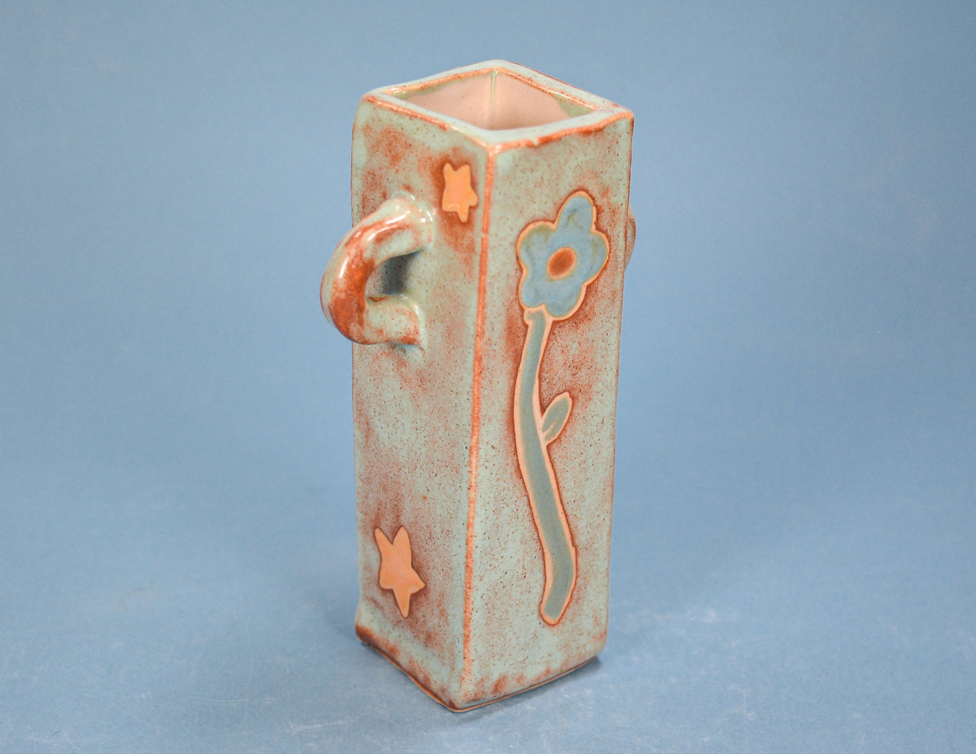 A small rectangular ceramic vase with a handle, featuring a light blue-green glaze with orange-brown distressed accents, a flower motif, and star shapes, set against a solid blue-gray background.