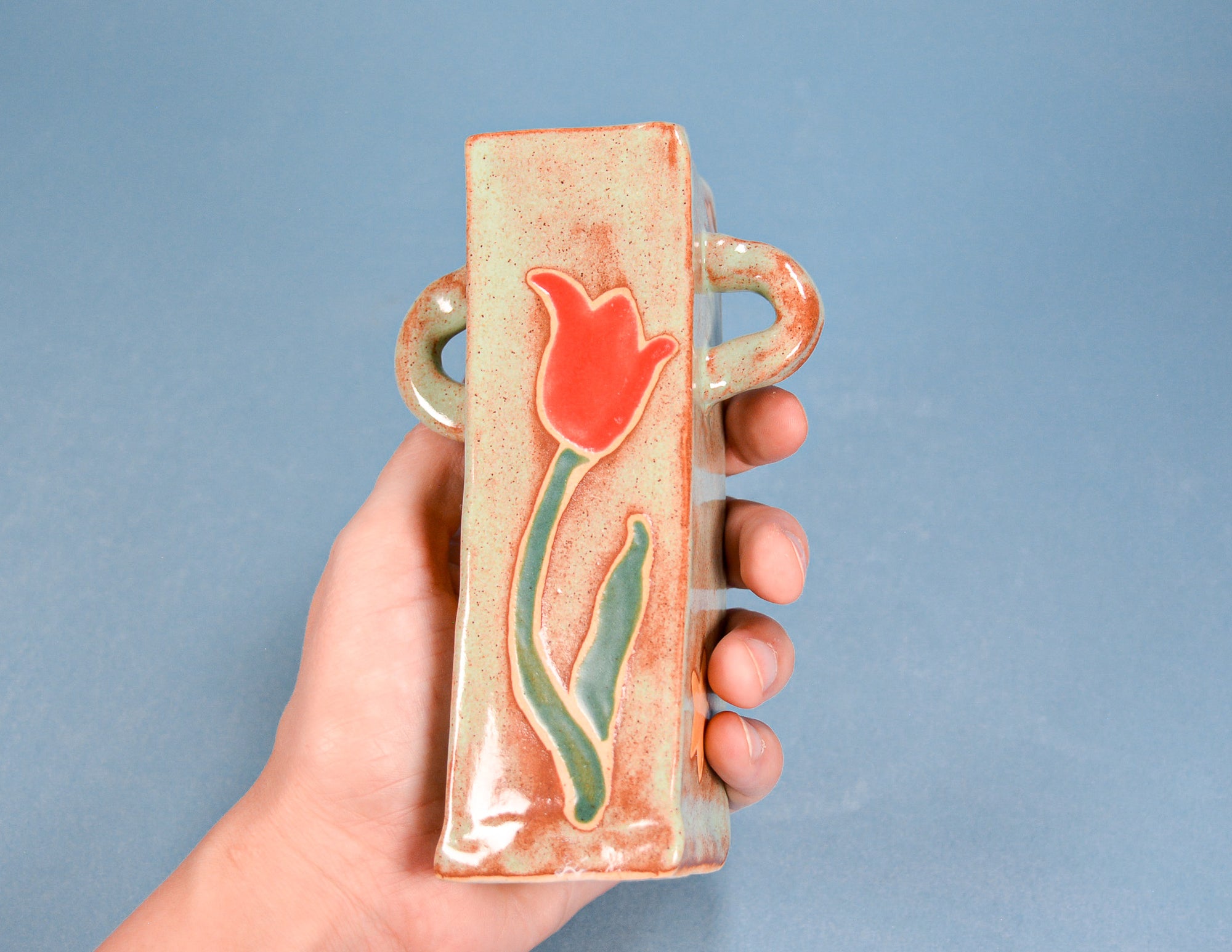 A hand holds a small rectangular ceramic vase with a raised red tulip design and green stem details against a solid blue background.
