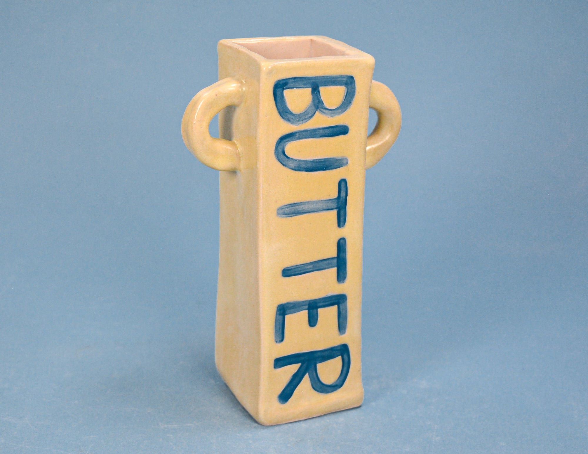 A beige ceramic butter dish with two side handles and the word 'BUTTER' painted vertically in blue on the front, set against a solid blue background.