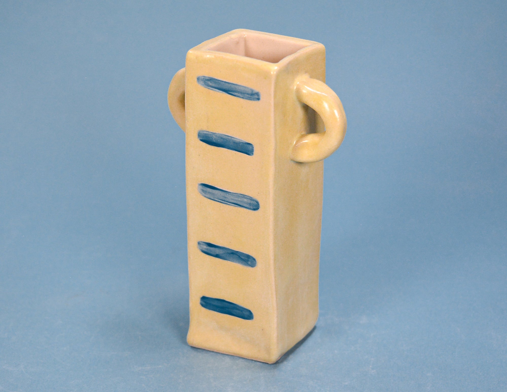 A small beige ceramic vase with a square open top, two loop handles on the sides, and five blue painted horizontal stripes down the front, set against a solid blue background.