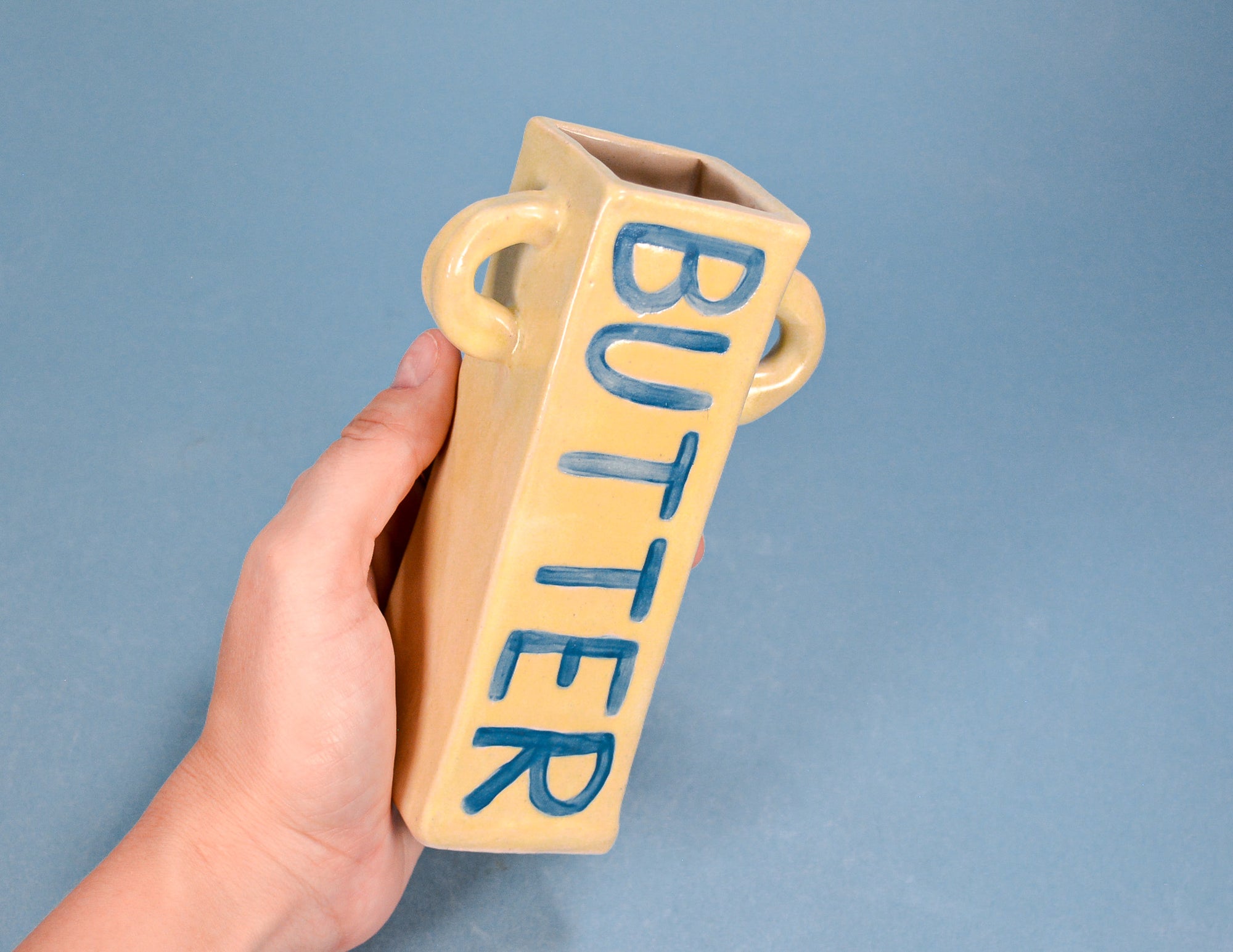 A hand holding a beige ceramic butter pail-shaped container with blue 'BUTTER' lettering, set against a solid blue background.