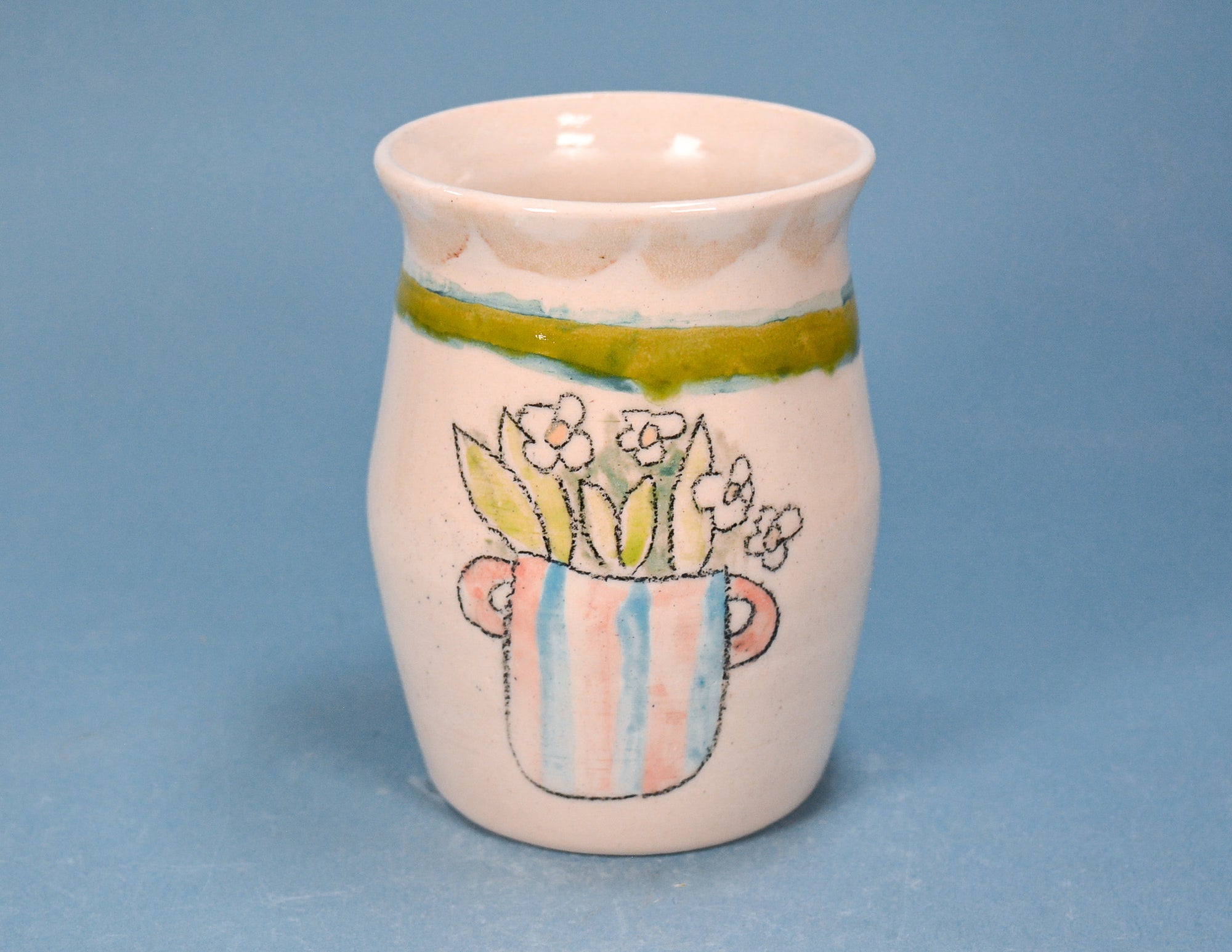 A small handmade ceramic cup with a flared rim, decorated with a painted floral design in a striped pot and a green band near the top, set against a solid blue background.