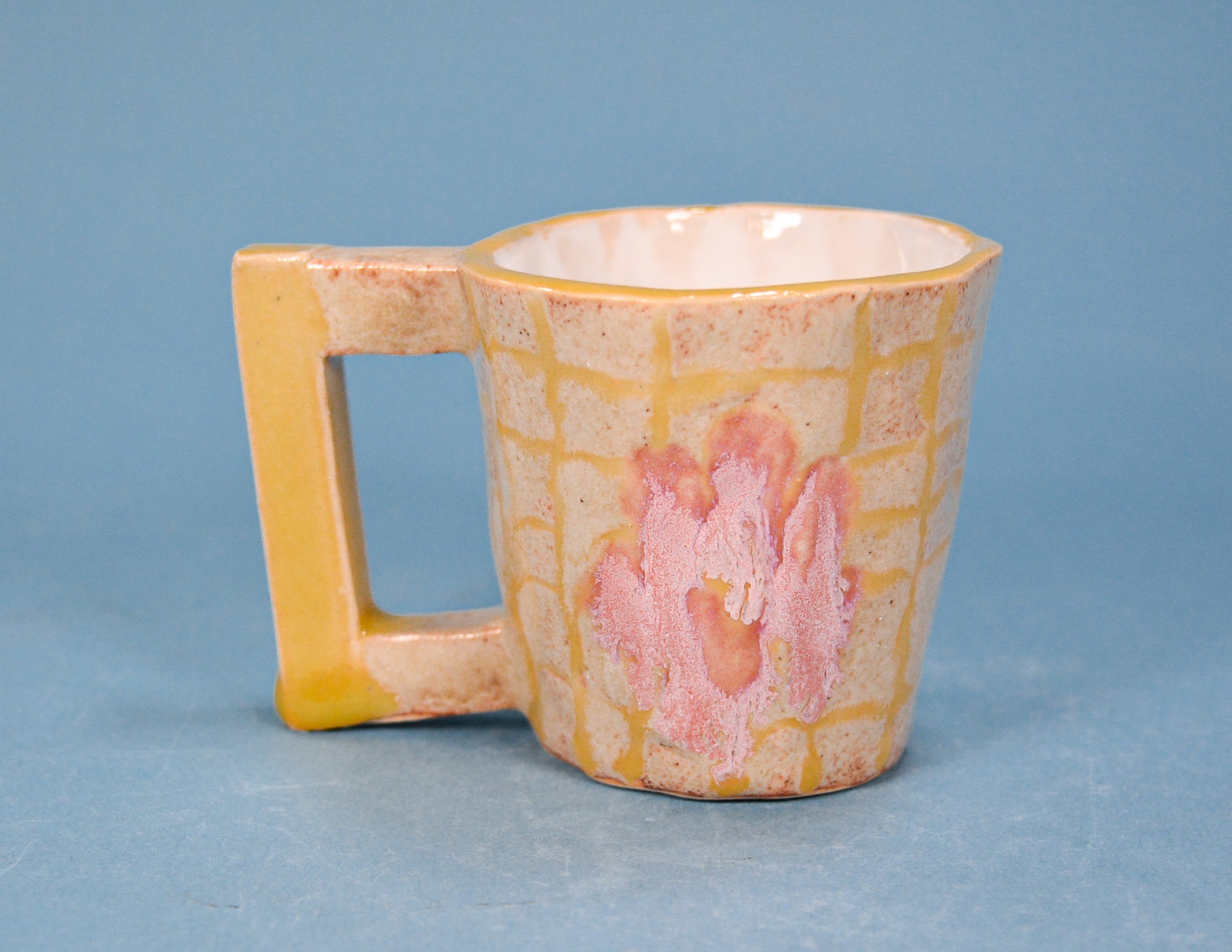 A small ceramic mug with a square handle, featuring a yellow grid pattern and a pink abstract floral design on a speckled beige base, set against a solid blue background.