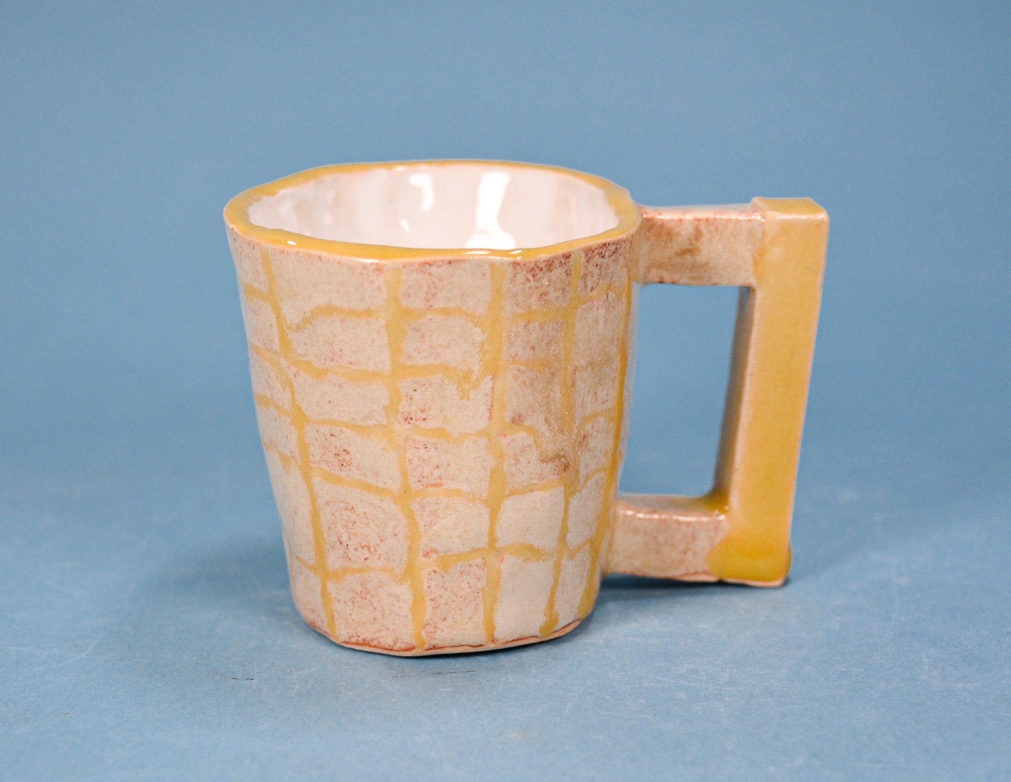 A handmade ceramic mug with a textured, grid-like pattern in cream and ochre yellow, featuring a thick rectangular handle, set against a solid blue background.