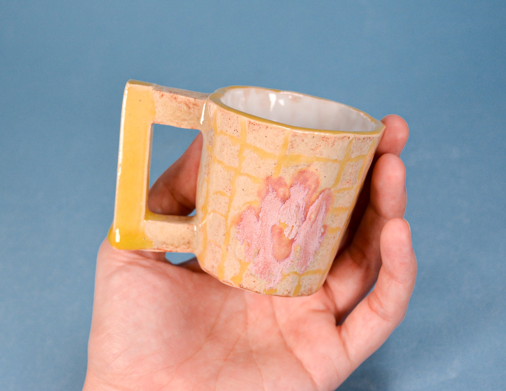 A hand holding a small ceramic mug with a yellow handle and a pink and yellow grid pattern on a blue background.