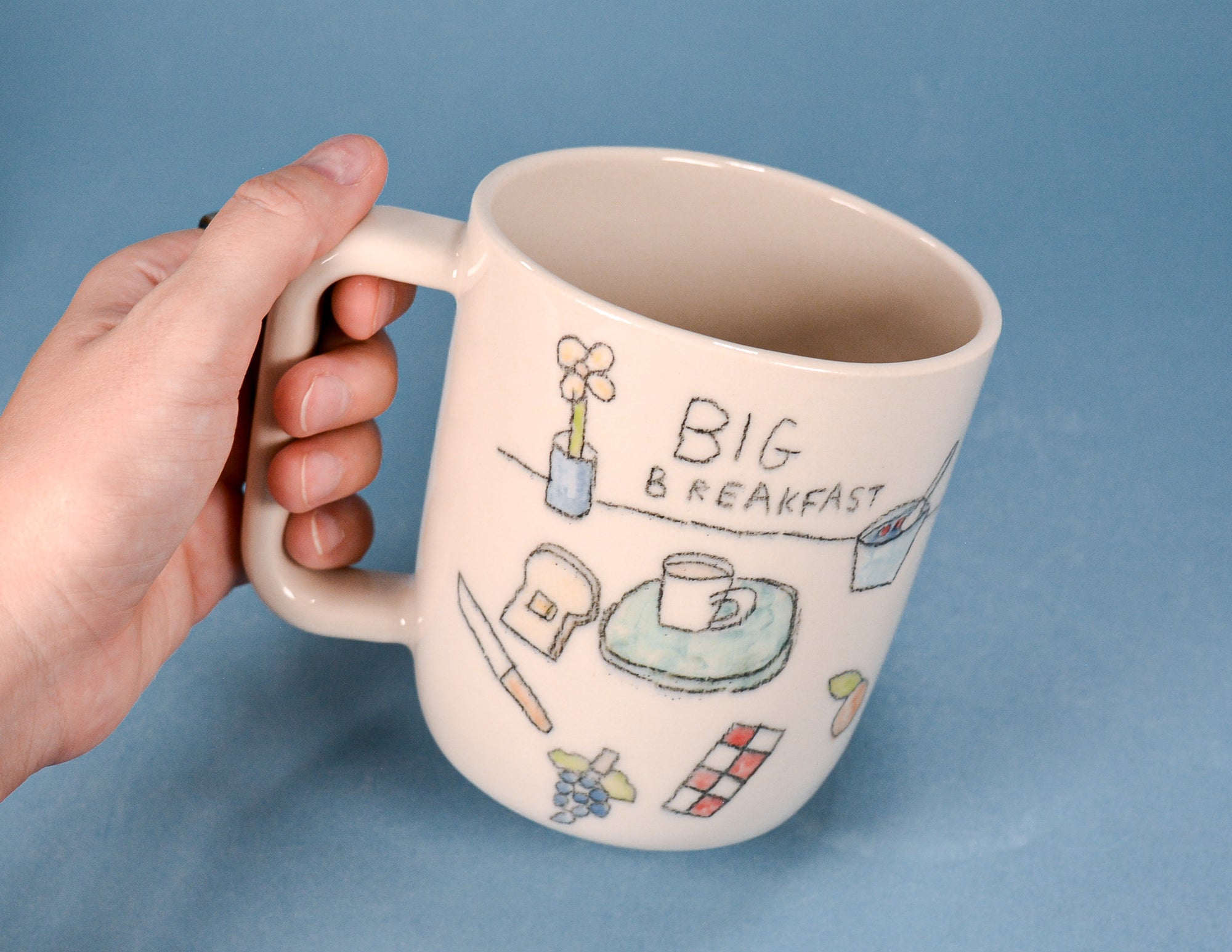 A hand holding a cream-colored ceramic mug with hand-drawn breakfast illustrations and the text 'BIG BREAKFAST' on a solid blue background.