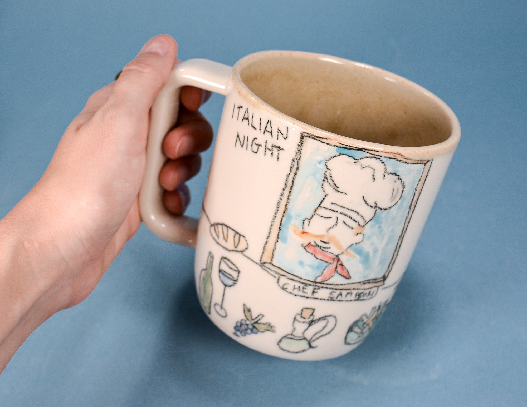 A hand holding a ceramic mug with a hand-painted illustration of Chef Carbonari and the text 'ITALIAN NIGHT' and 'CHEF CARBONARI' against a solid blue background.