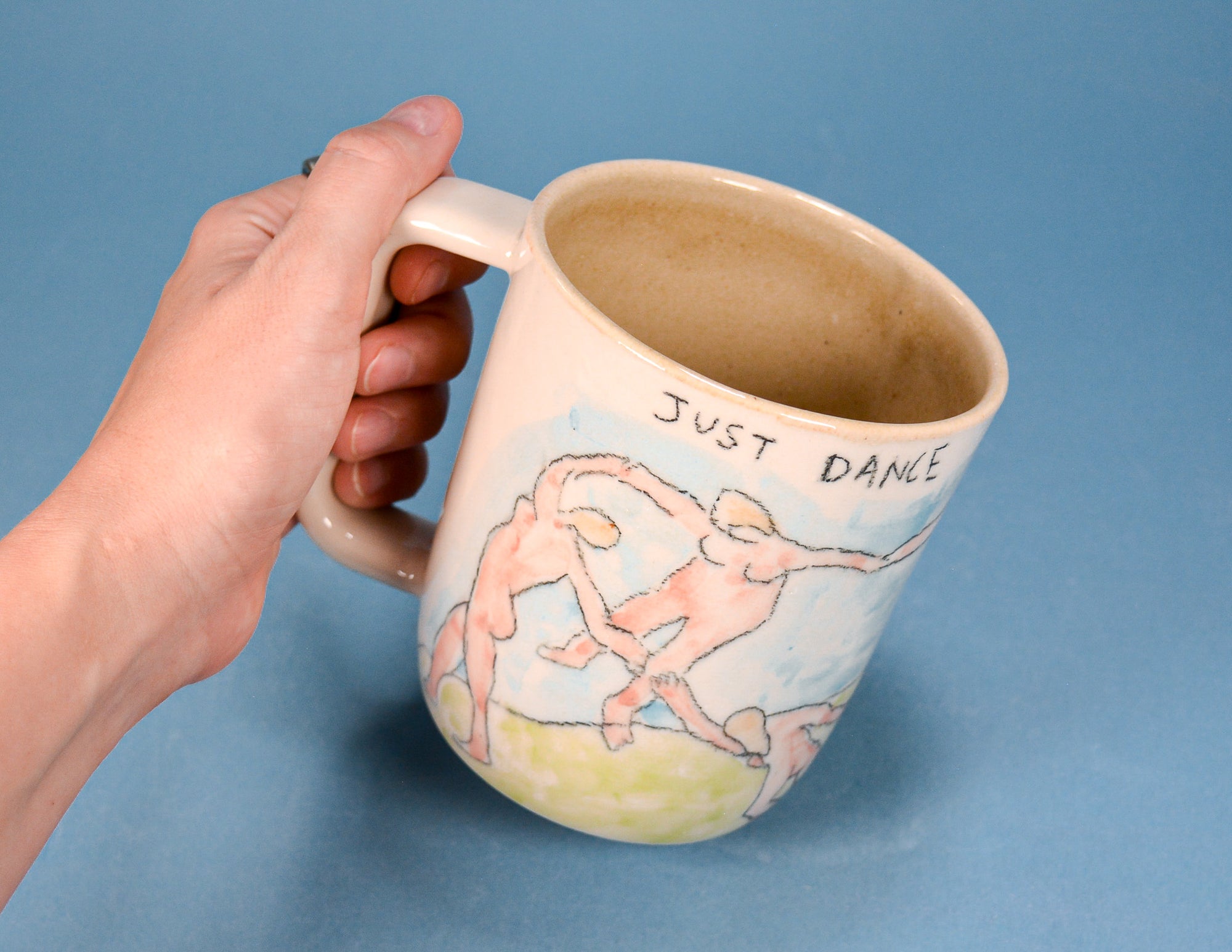A hand holding a ceramic mug with a hand-drawn illustration of two dancing figures and the text 'JUST DANCE' on a solid blue background.