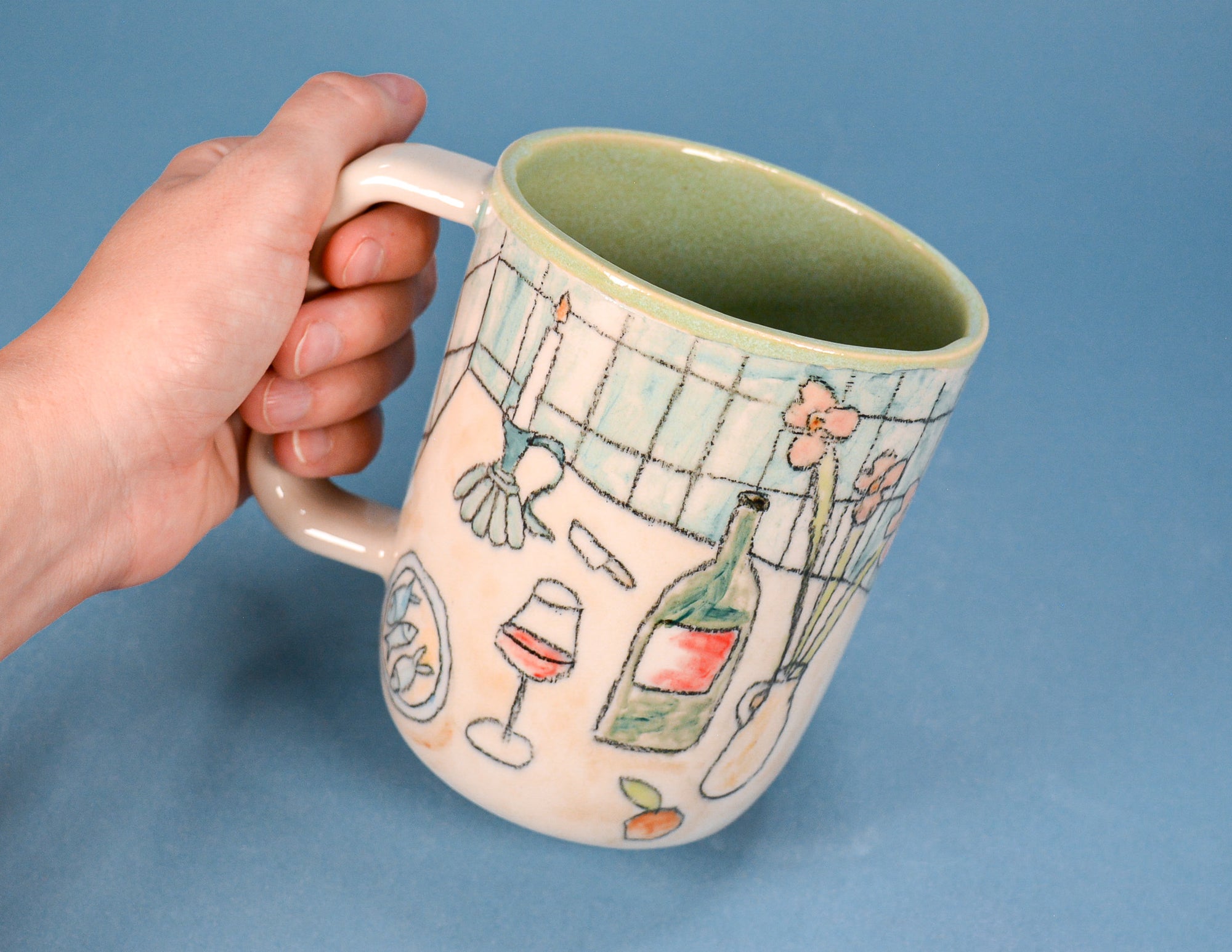 A hand holding a ceramic mug with a light pink exterior decorated with hand-drawn illustrations of kitchen and dining items, and a light green interior, set against a solid blue background.