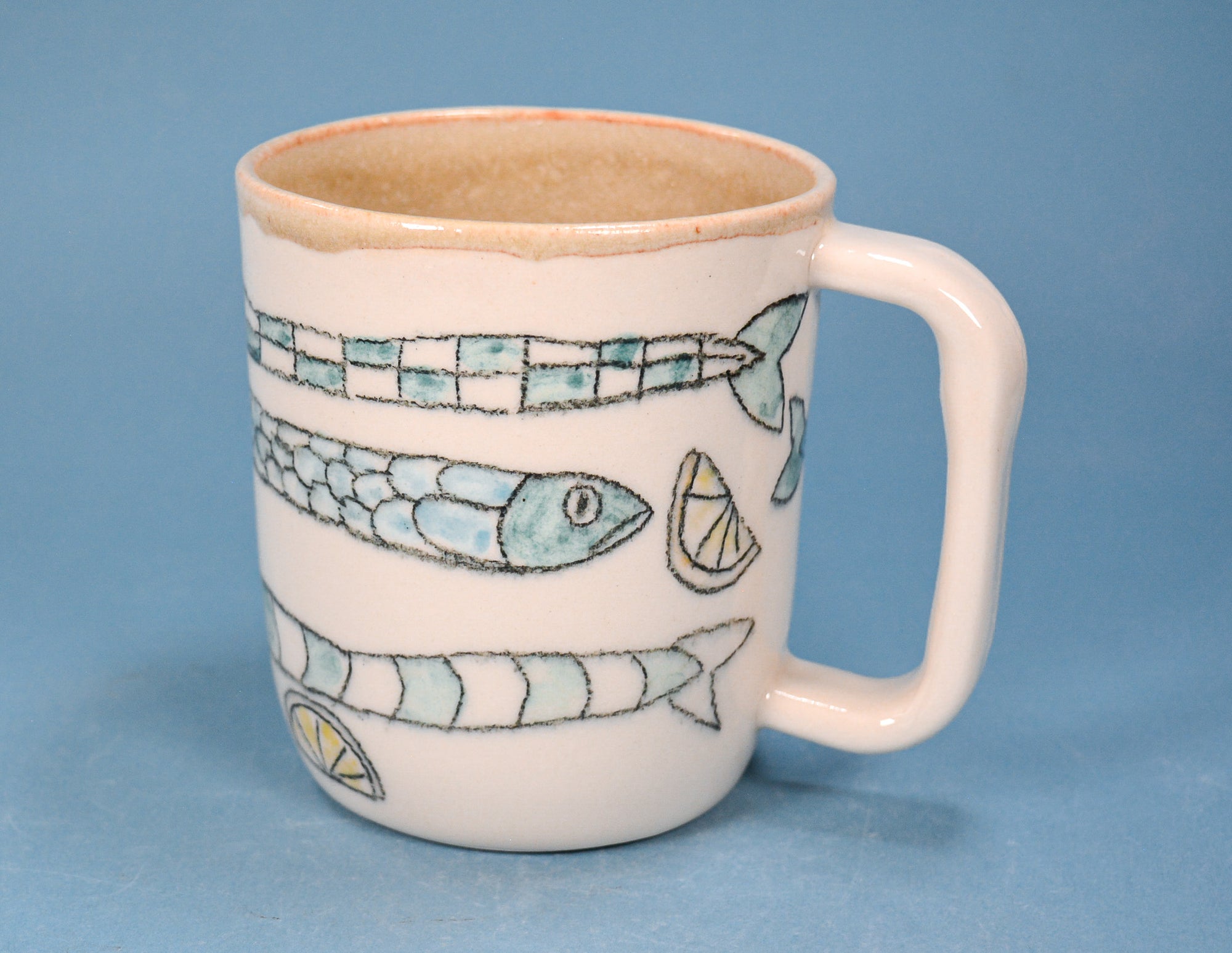 A white ceramic mug with a handle, decorated with hand-painted fish and lemon slice motifs in light blue and green, set against a solid blue background.