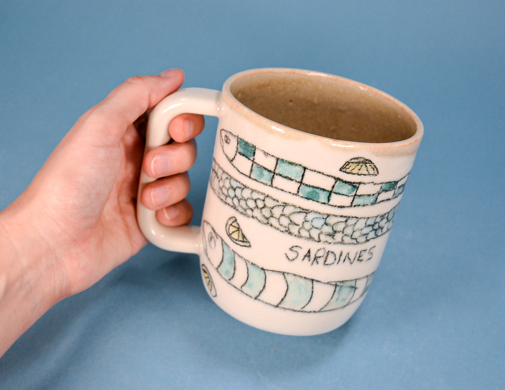 A hand holding a ceramic mug with a blue and white sardine pattern and the word 'SARDINES' printed on it, set against a solid blue background.