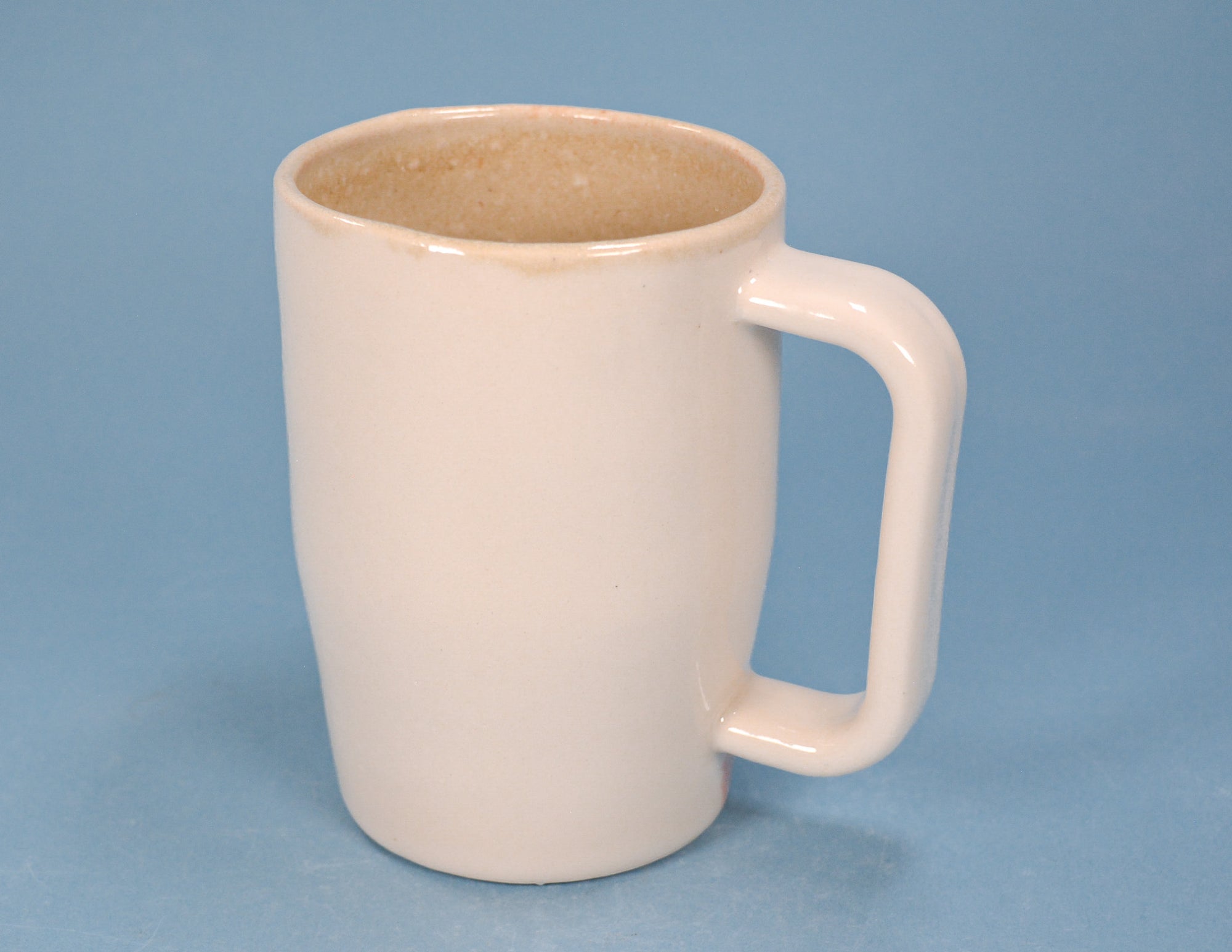 A ceramic mug with a rounded body and a thick, integrated handle, featuring a matte off-white exterior and a speckled beige interior, set against a solid blue background.