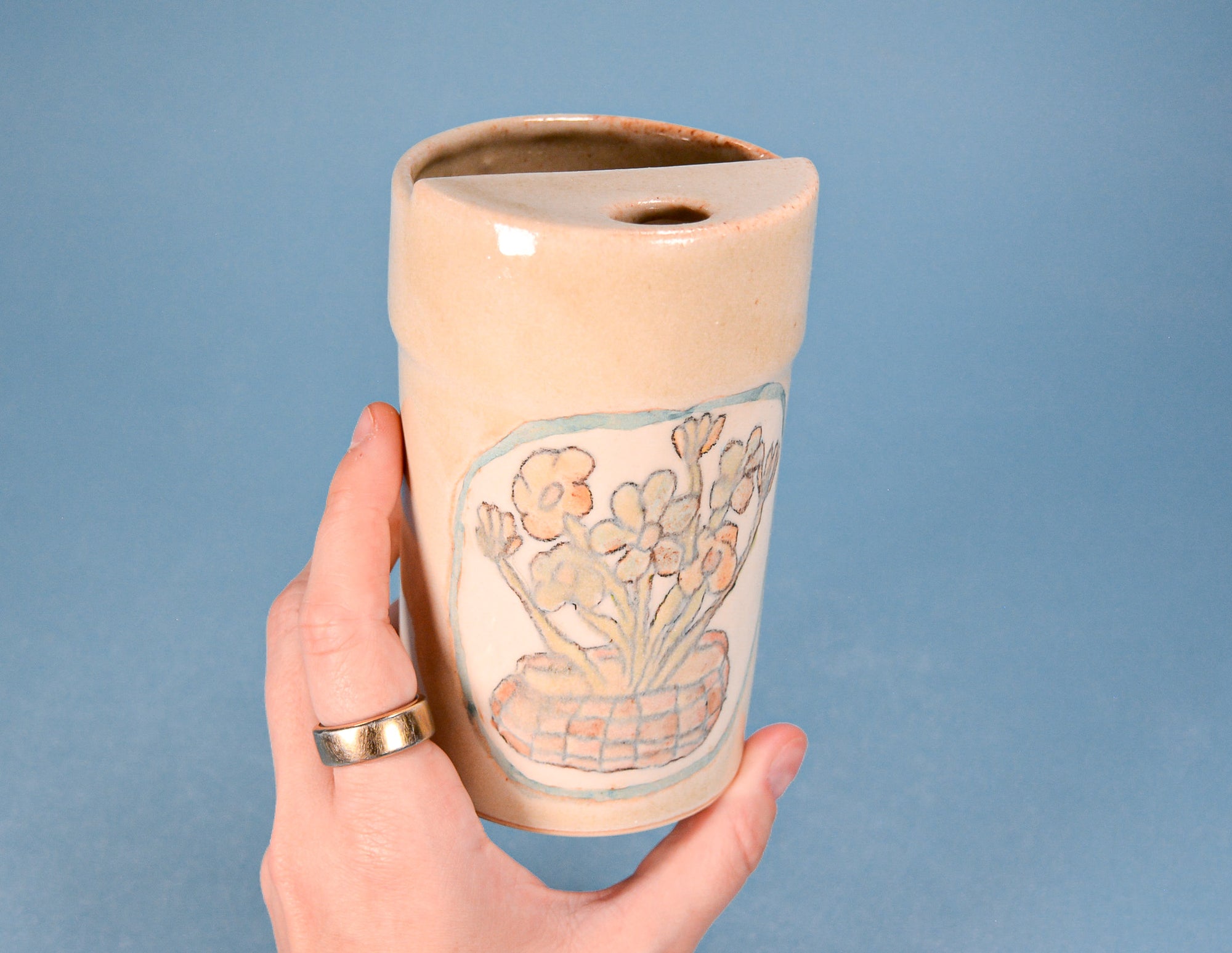 A hand holding a small handmade ceramic planter with a floral illustration, set against a solid blue background.