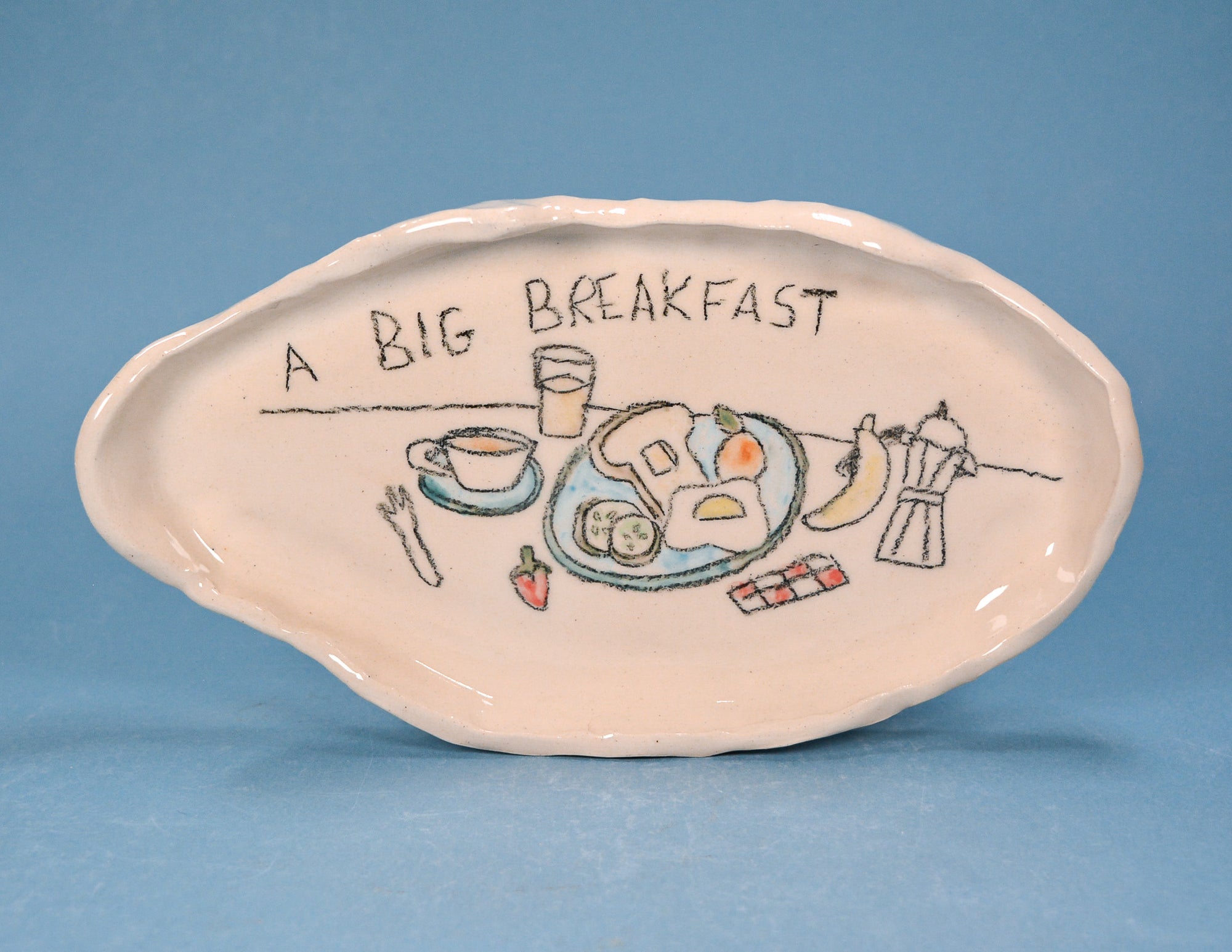 An off-white ceramic breakfast plate with a hand-drawn illustration of a breakfast table and the text 'A BIG BREAKFAST' written on it, set against a solid blue background.