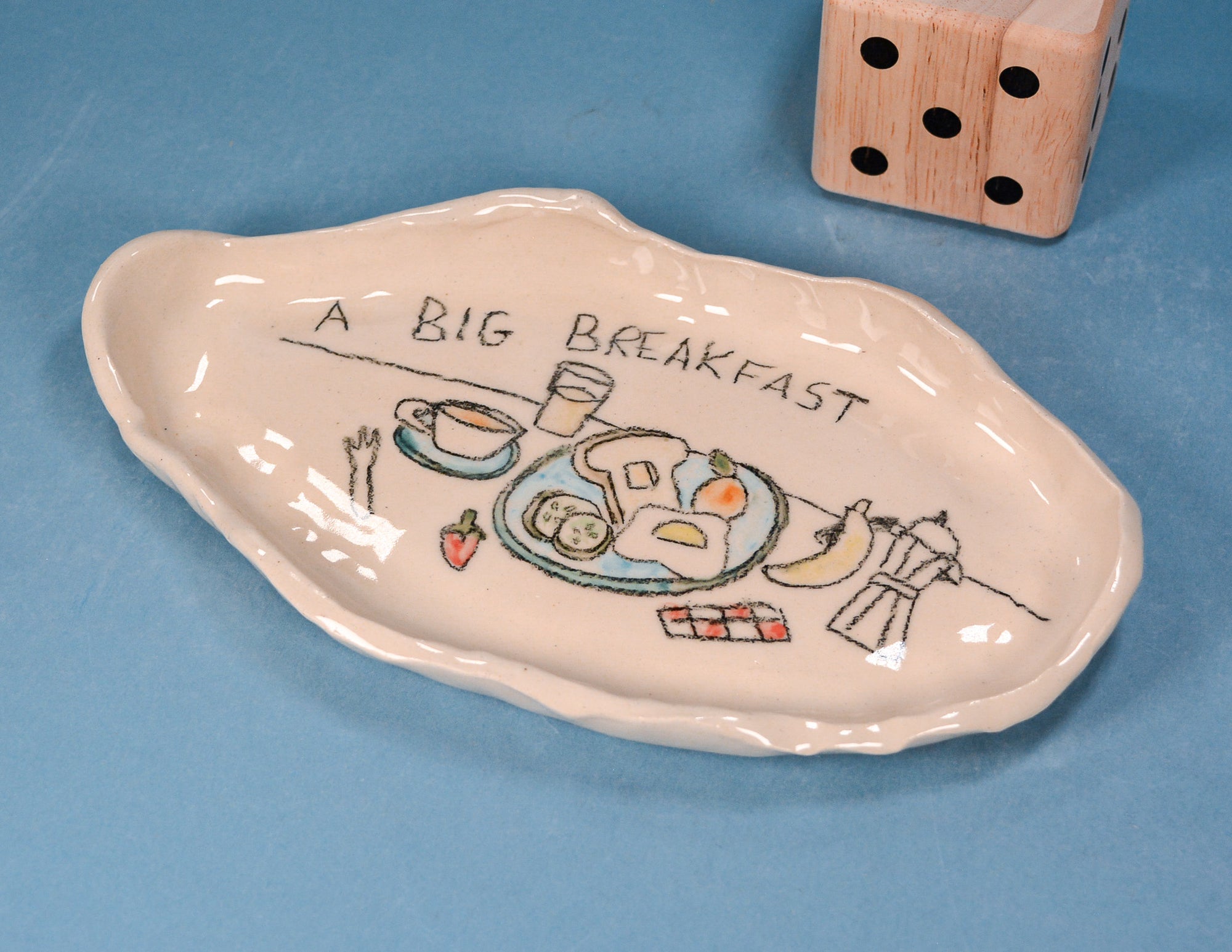 An irregularly shaped ceramic breakfast plate with a hand-drawn illustration of a meal and the text 'A BIG BREAKFAST' on a blue background, with a wooden die partially visible in the top right corner.