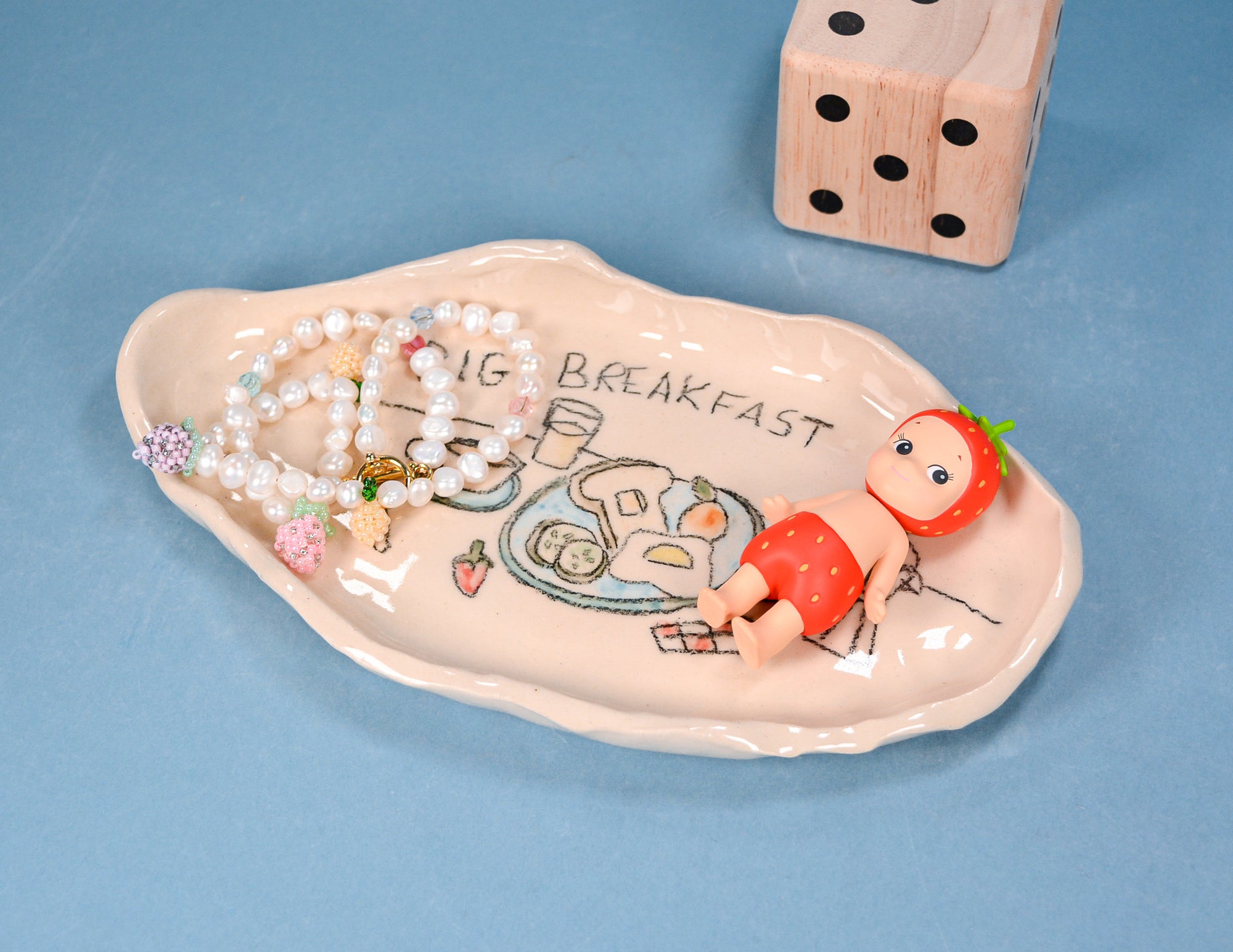 A ceramic breakfast-themed dish with a cartoon character and the words 'BIG BREAKFAST' on a light blue background, styled with pearl beads and a wooden die.
