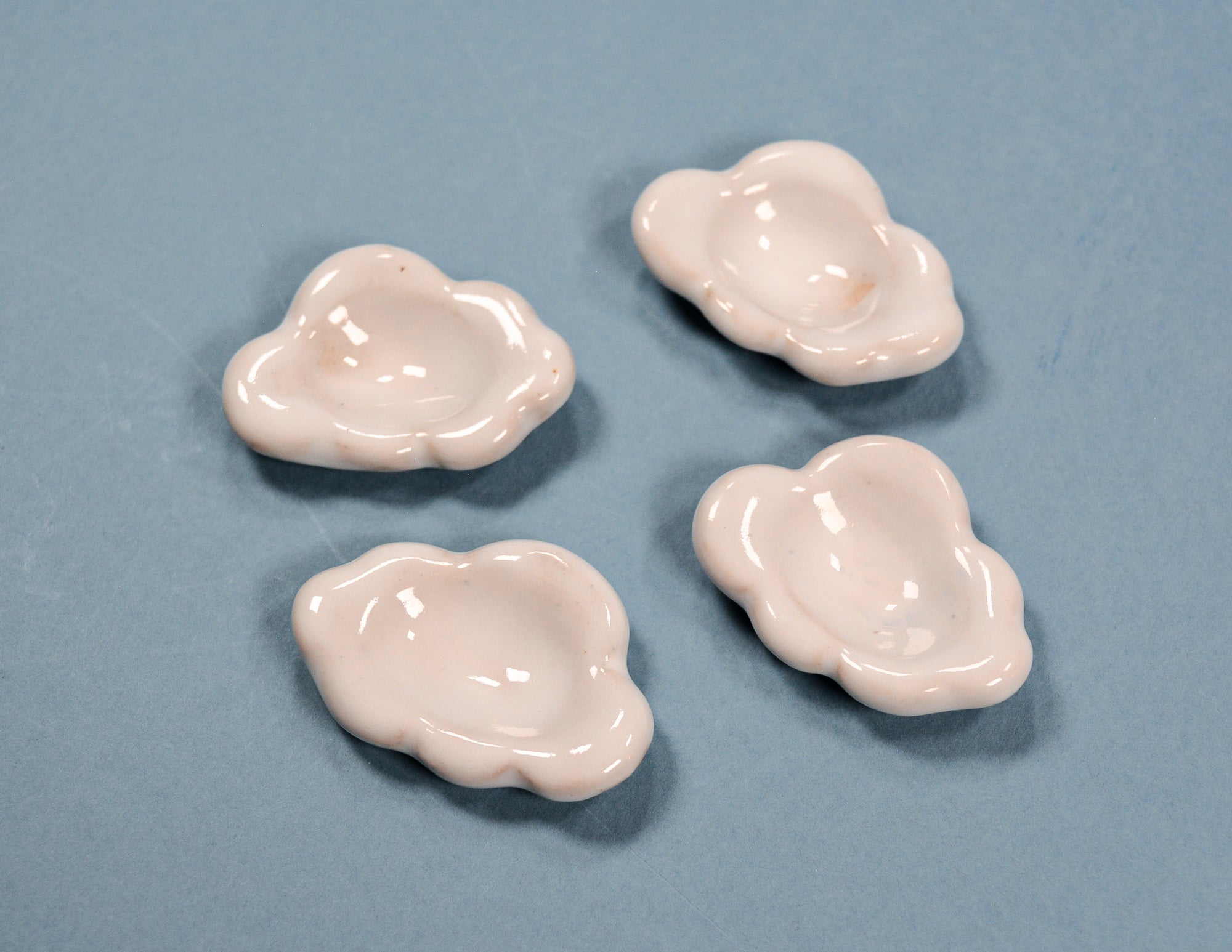 Four small glossy cloud-shaped ceramic dish holders in a pale pink color, arranged on a solid blue-gray background.