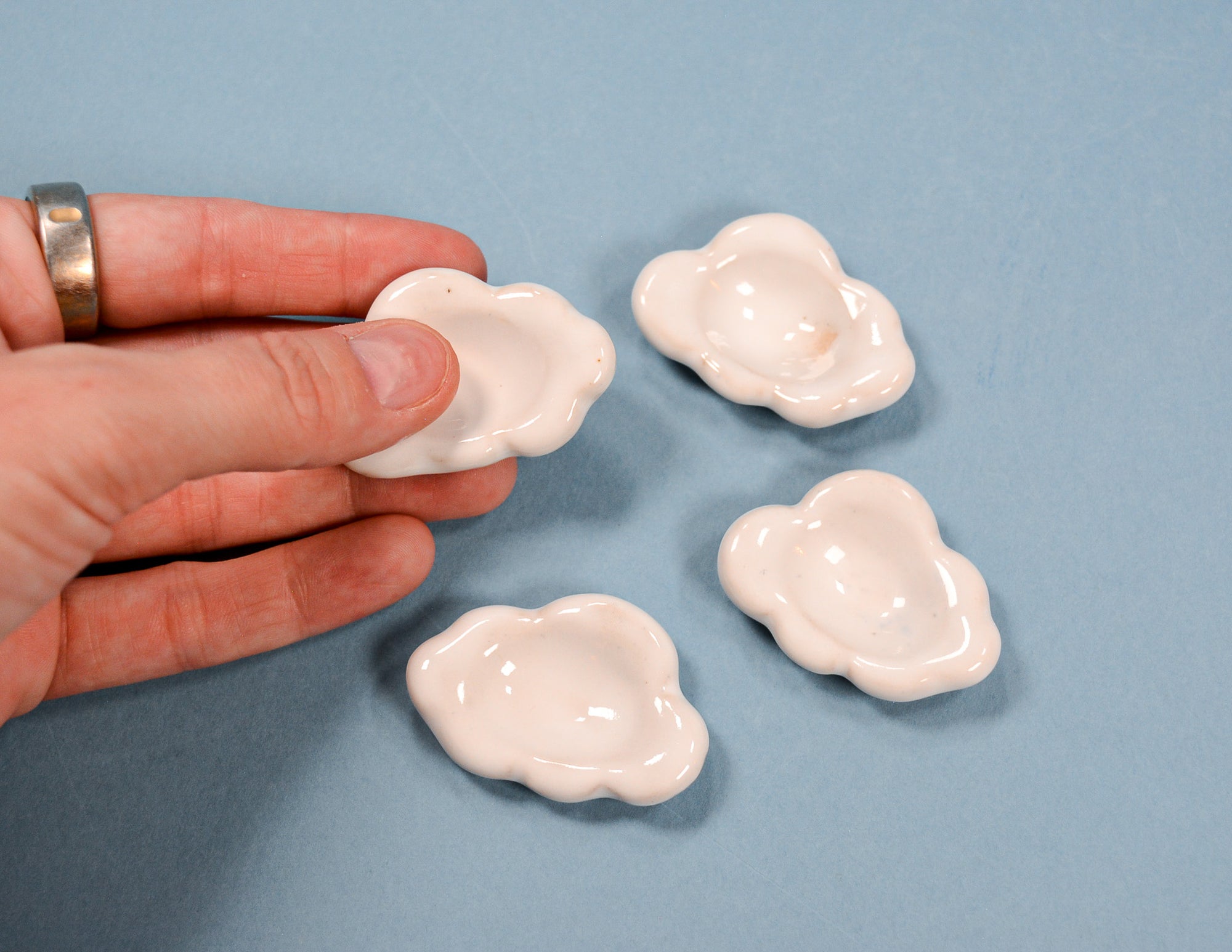Four small glossy white ceramic dish holders with a wavy, cloud-like outline, with one being held by a hand to show scale against a solid blue background.
