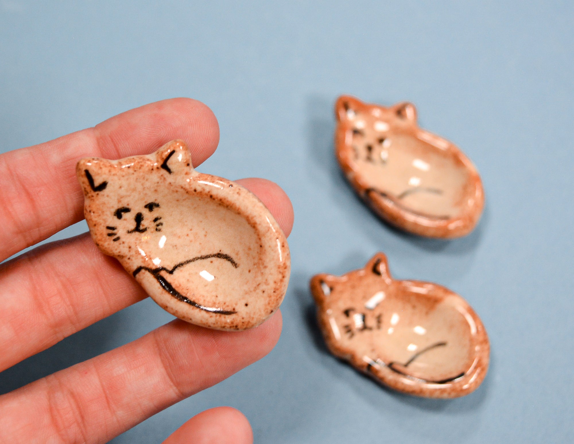 Three small ceramic cat-shaped bowls with a speckled beige and brown glaze are shown, with one held in a person's hand and two resting on a light blue background.