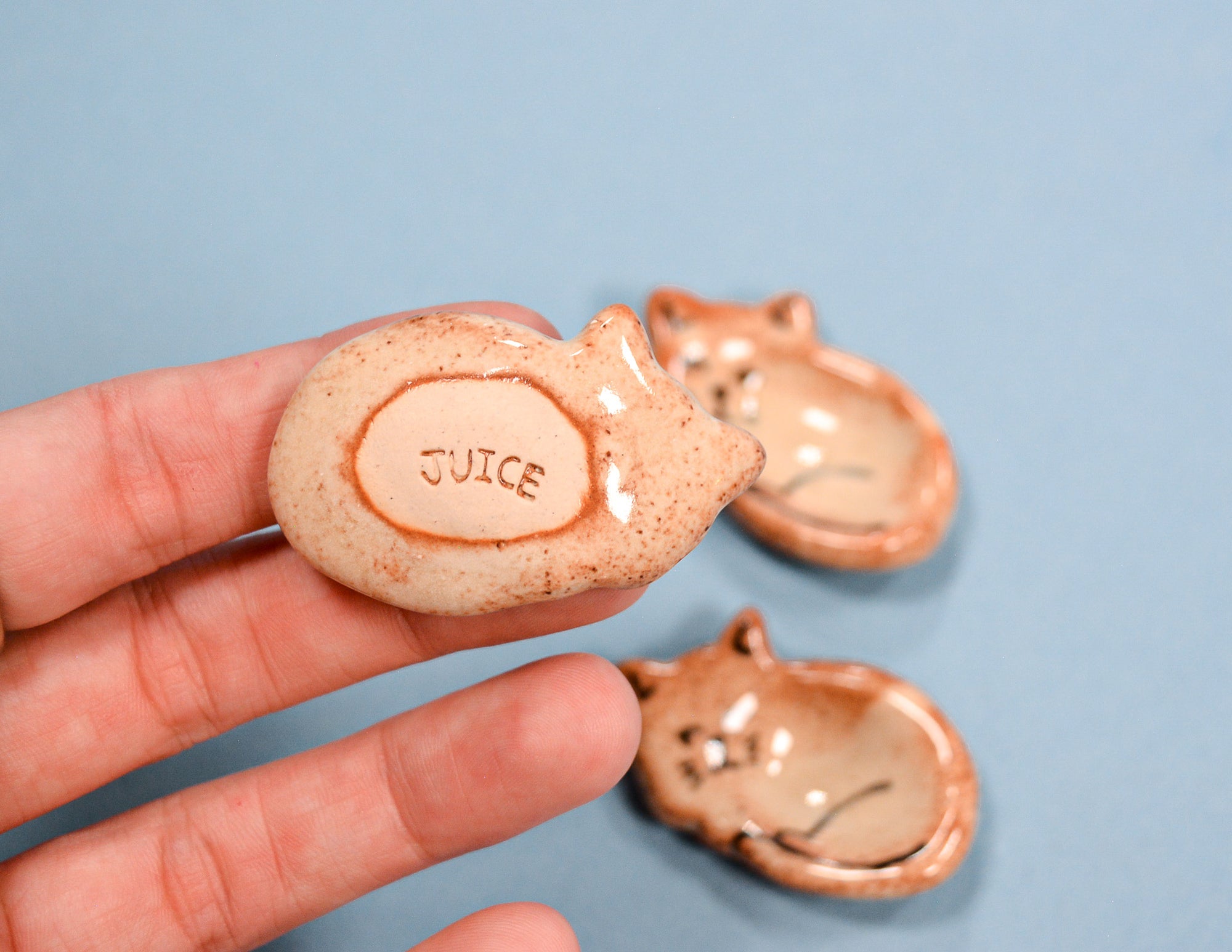 A hand holds a small ceramic cat-shaped juice cup with the word 'JUICE' molded into the front, with two more cat-shaped juice cups resting on a light blue background.