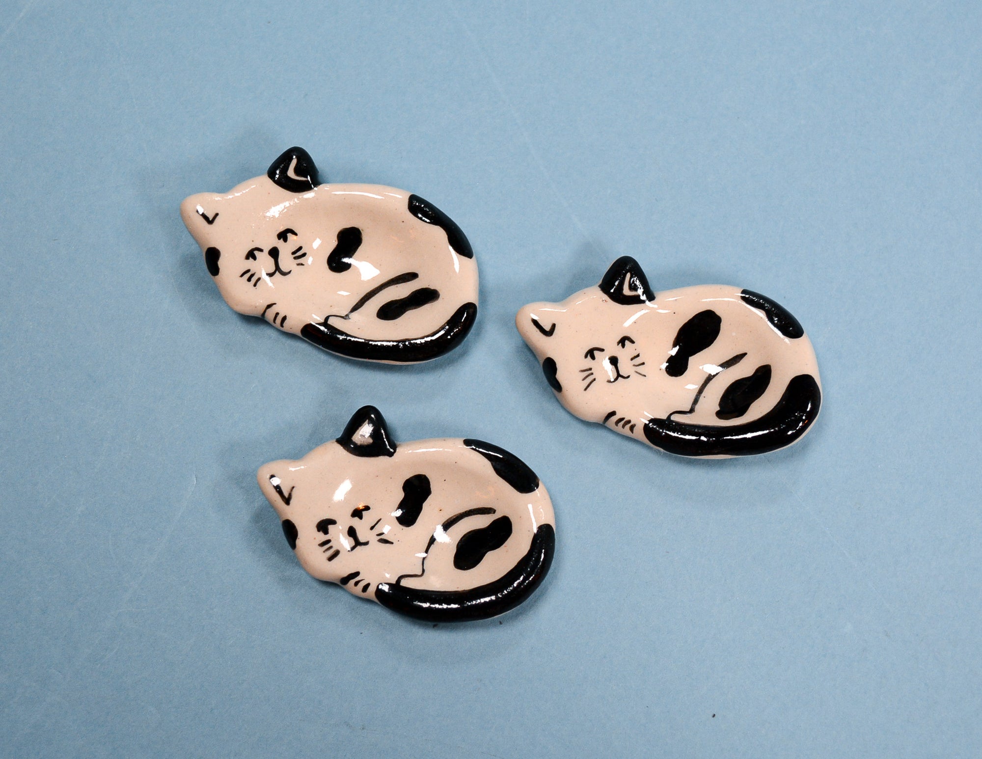 Three small ceramic cat-shaped dishes with a glossy finish, featuring a light pink body with black spots and black outlines, arranged on a solid light blue background.
