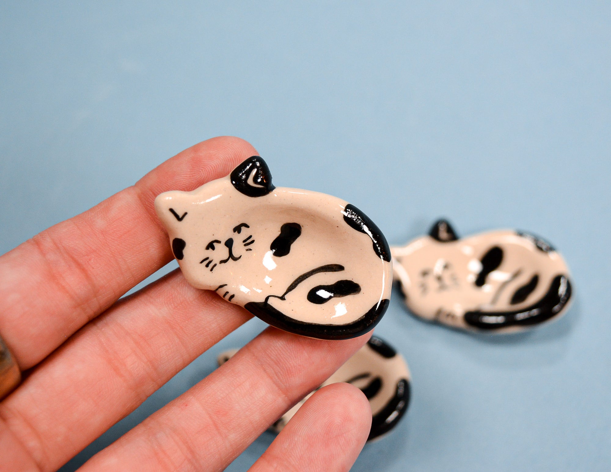 A hand holding a small ceramic cat-shaped dish with a cream body and black painted markings, with two more similar dishes blurred in the background on a light blue surface.