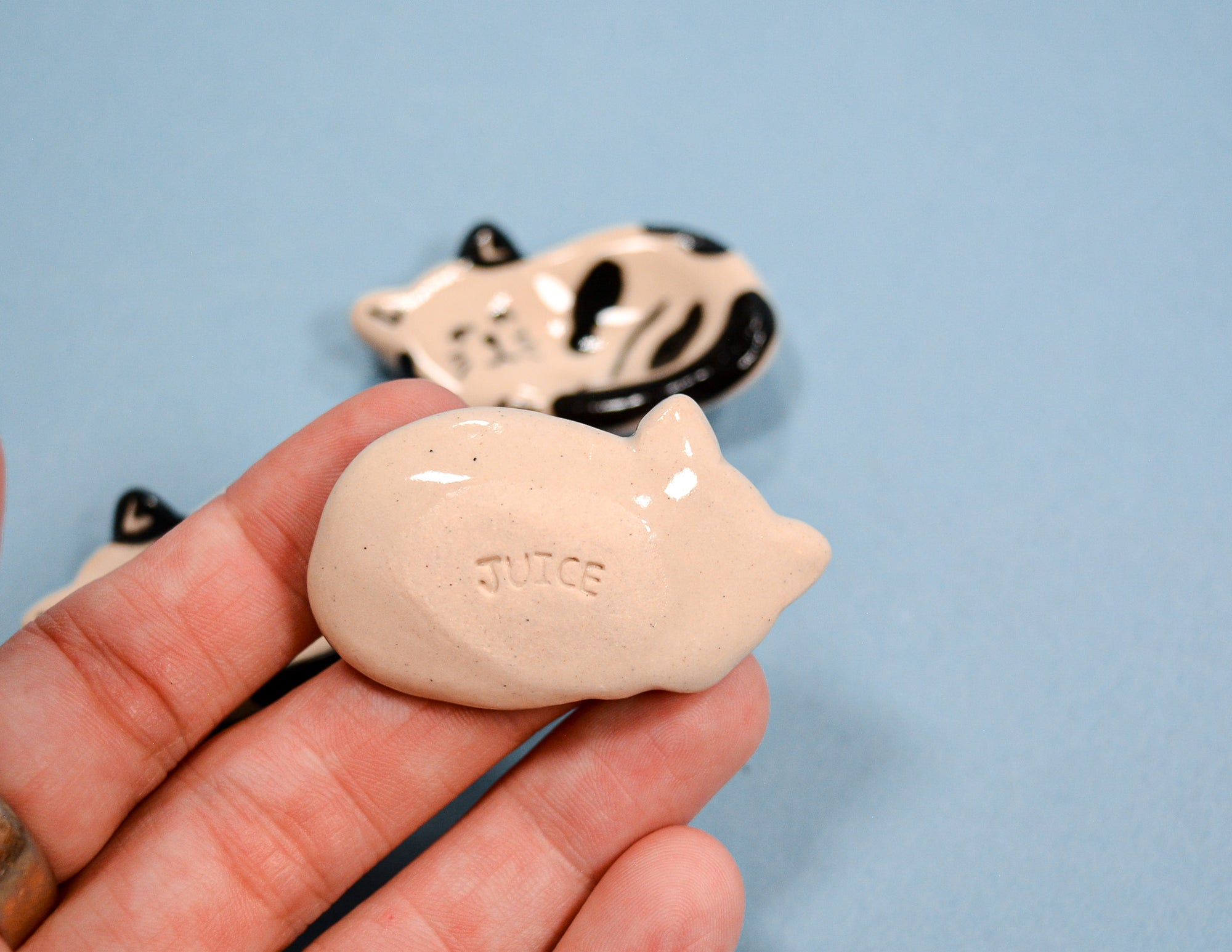A hand holding a small glossy ceramic pig-shaped object with the word 'JUICE' molded into it, with two more similar pig-shaped items blurred in the background against a light blue backdrop.
