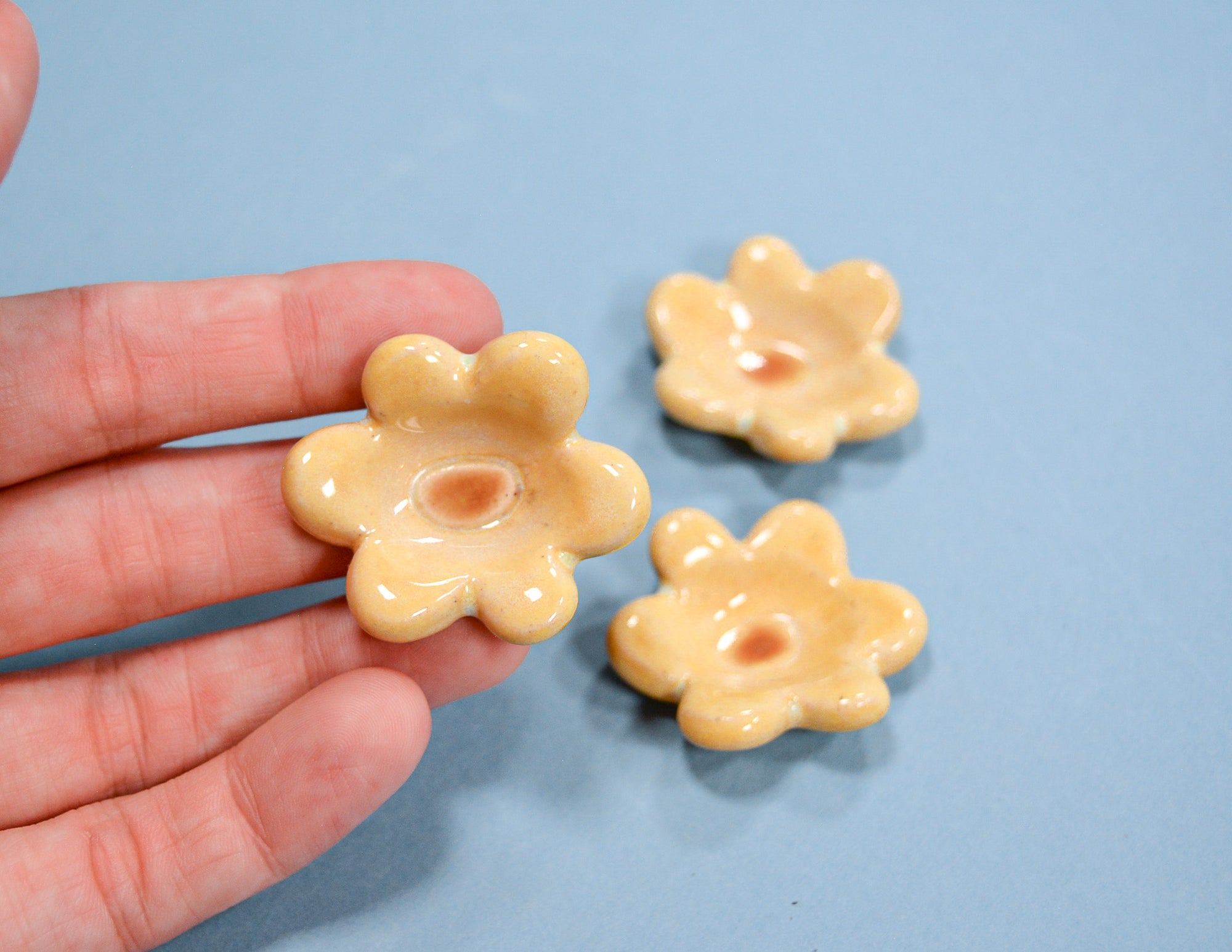 Three small, glossy, flower-shaped beads in a pale yellowish-beige color are shown, with one held in a person's hand and two resting on a light blue background.