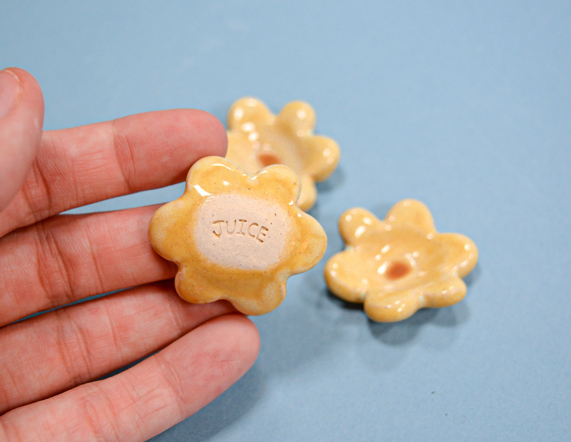 A hand holds a small flower-shaped candy with the word 'JUICE' embossed in the center, with two more of the same candies resting on a light blue surface in the background.