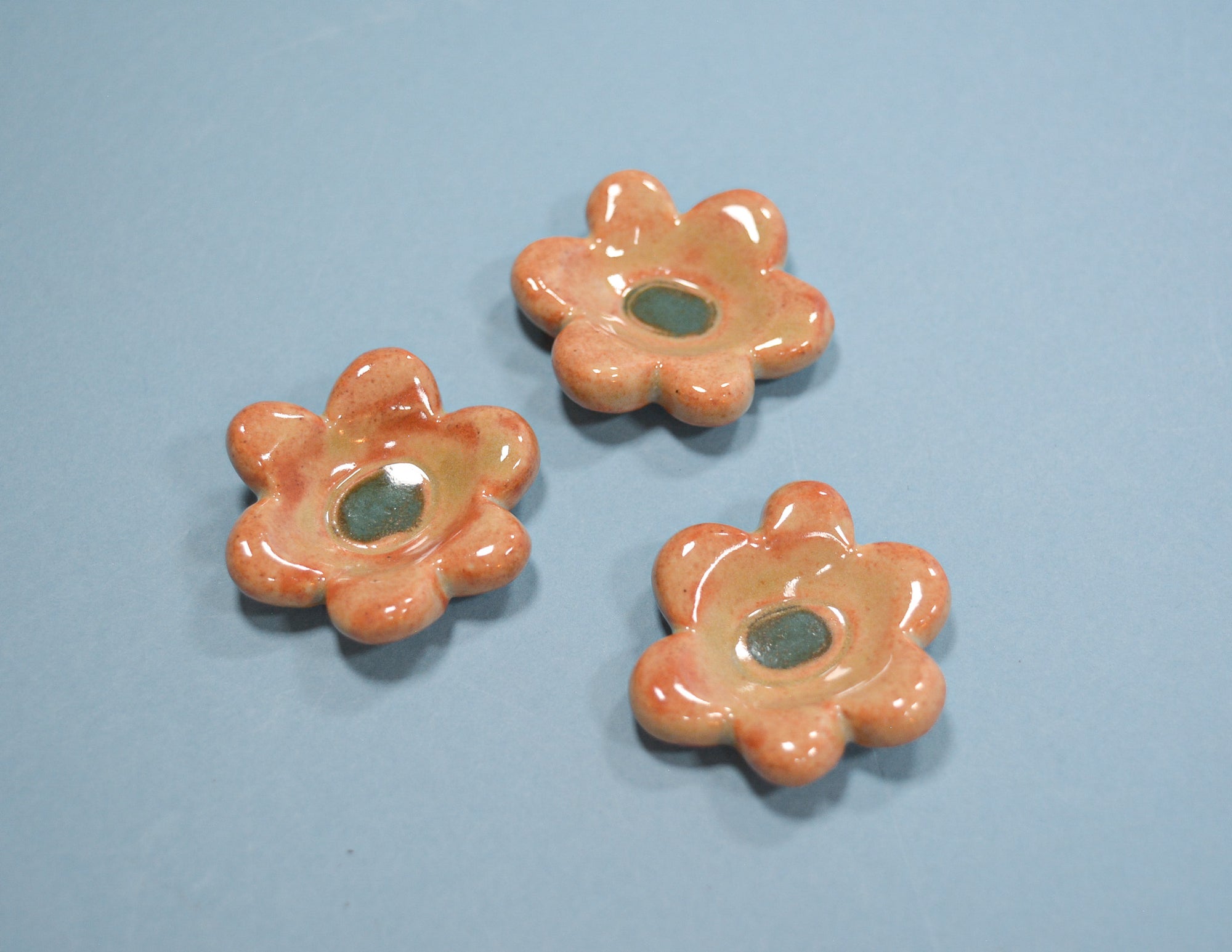Three small flower-shaped ceramic dish pieces in a glossy peach-orange glaze with a dark green center, arranged on a light blue background.
