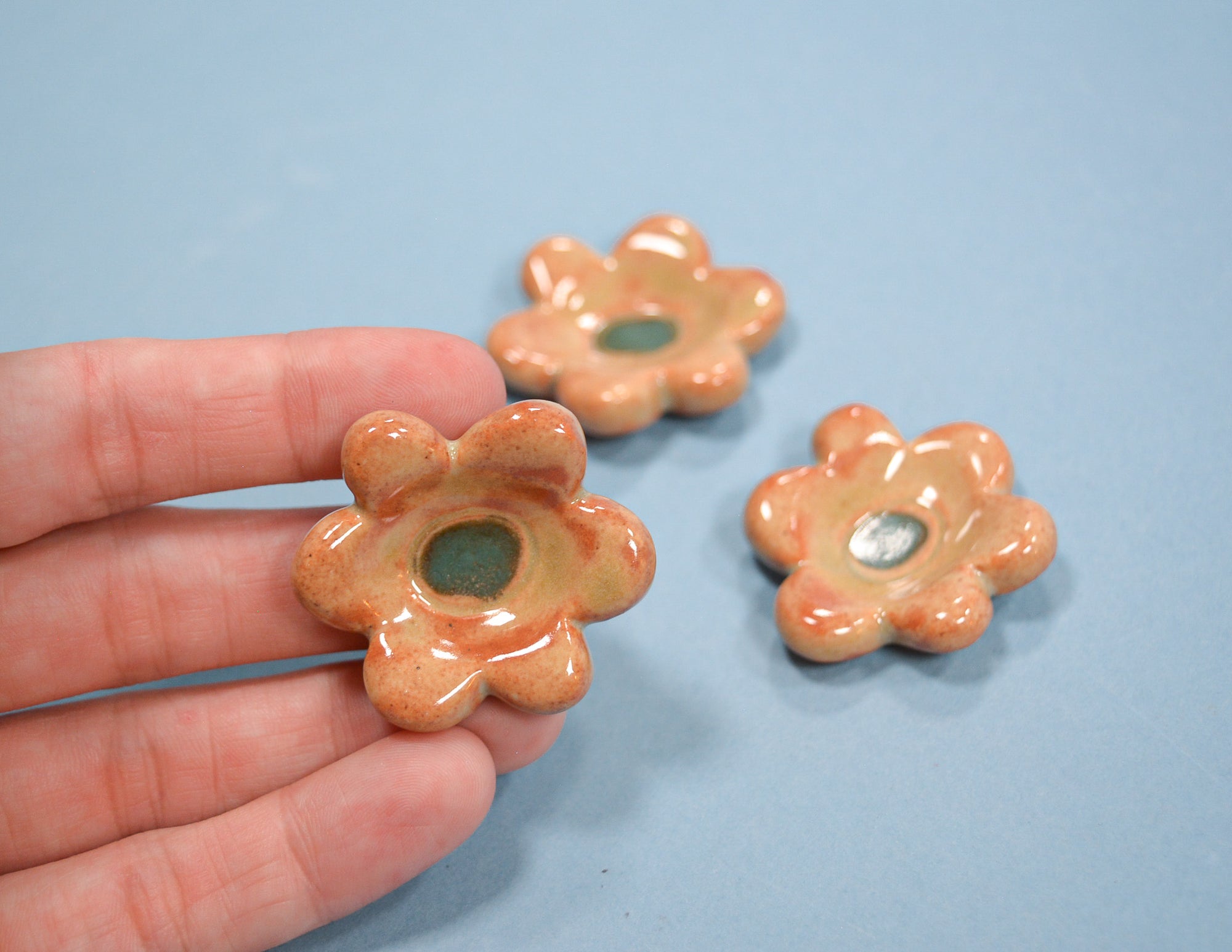 Three small flower-shaped ceramic pieces in a glossy peach-orange glaze with green centers, shown on a light blue background with one held in a hand for scale.