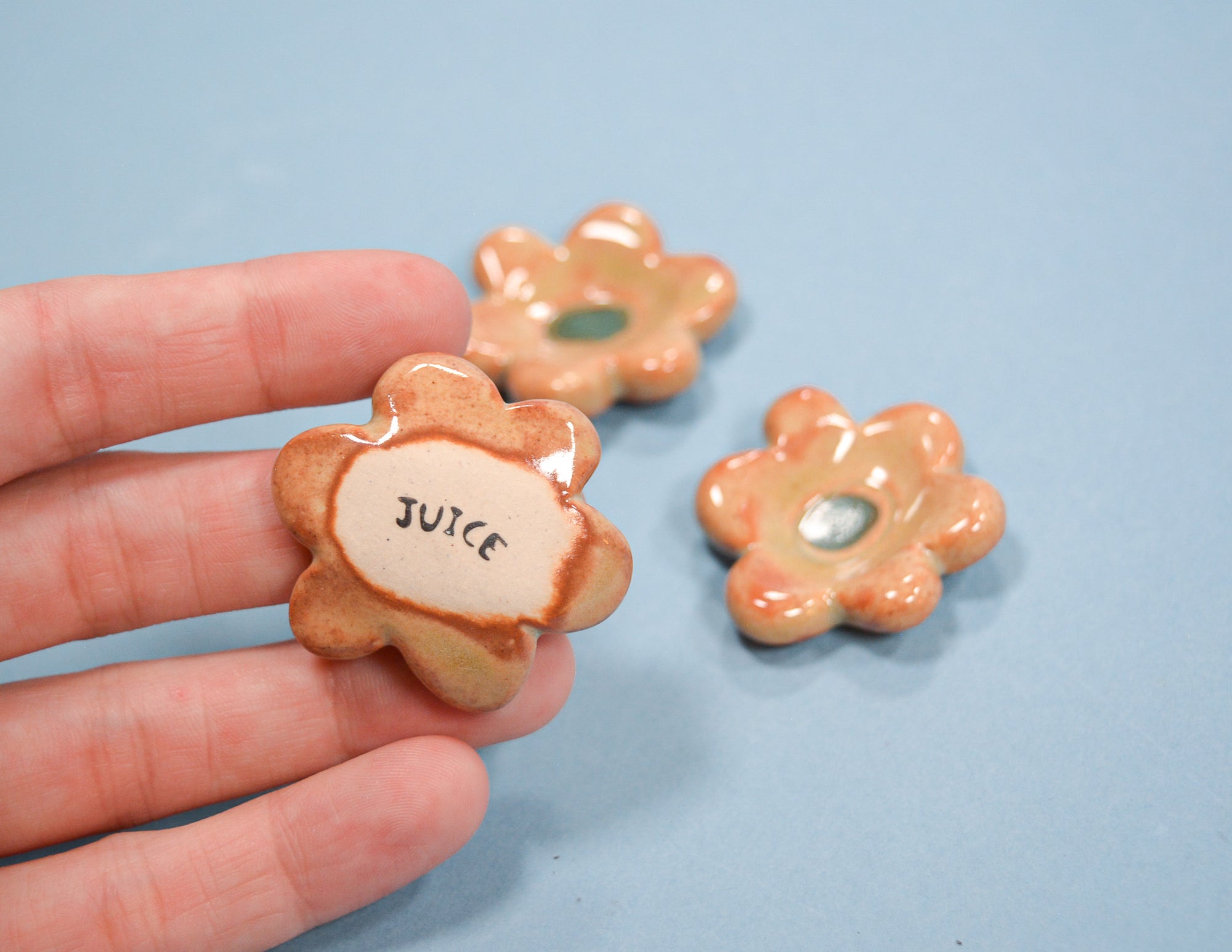 A hand holds a small flower-shaped ceramic cup labeled 'JUICE' with a glossy peach and beige glaze, while two matching cups sit on a light blue surface in the background.