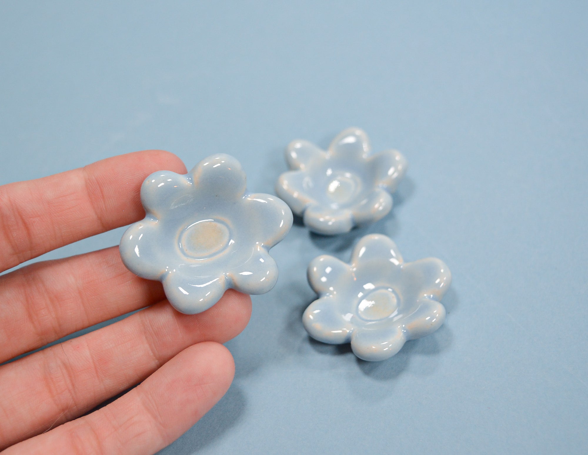 Three small glossy flower-shaped ceramic dishes in a pale icy blue color, with one held in a person's hand and two resting on a light blue surface.